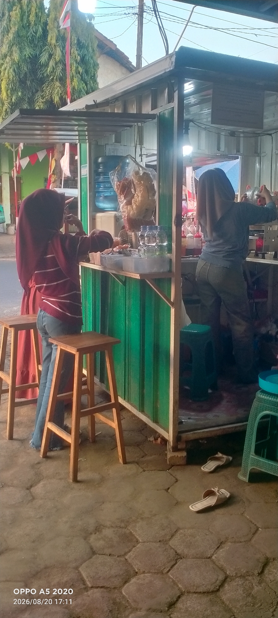 The image depicts an outdoor food stall located on a street in Kedung, Indonesia. The stall is constructed with green corrugated metal siding and has a white roof. A young woman wearing a dark red headscarf and a striped sweater is standing at the front of the stall, facing away from the camera. She is wearing blue jeans and appears to be interacting with items on the counter. To her left, another wooden stool is visible.
Behind her, a second woman, also wearing a headscarf and a light blue shirt, is working inside the stall. She is seen from the back, and her hands are reaching towards something on the counter. The stall's counter displays various items, including bottles of water, plastic containers, and bags of what appear to be snacks. A large blue water dispenser is also visible behind the counter.
The ground in front of the stall is paved with hexagonal concrete tiles. A pair of light-colored sandals are lying on the ground to the right of the stall. In the background, there are trees with red and white Indonesian flags, suggesting a festive or commemorative occasion. Power lines are visible stretching across the sky. The overall scene is consistent with a typical street-side food vendor in Indonesia.