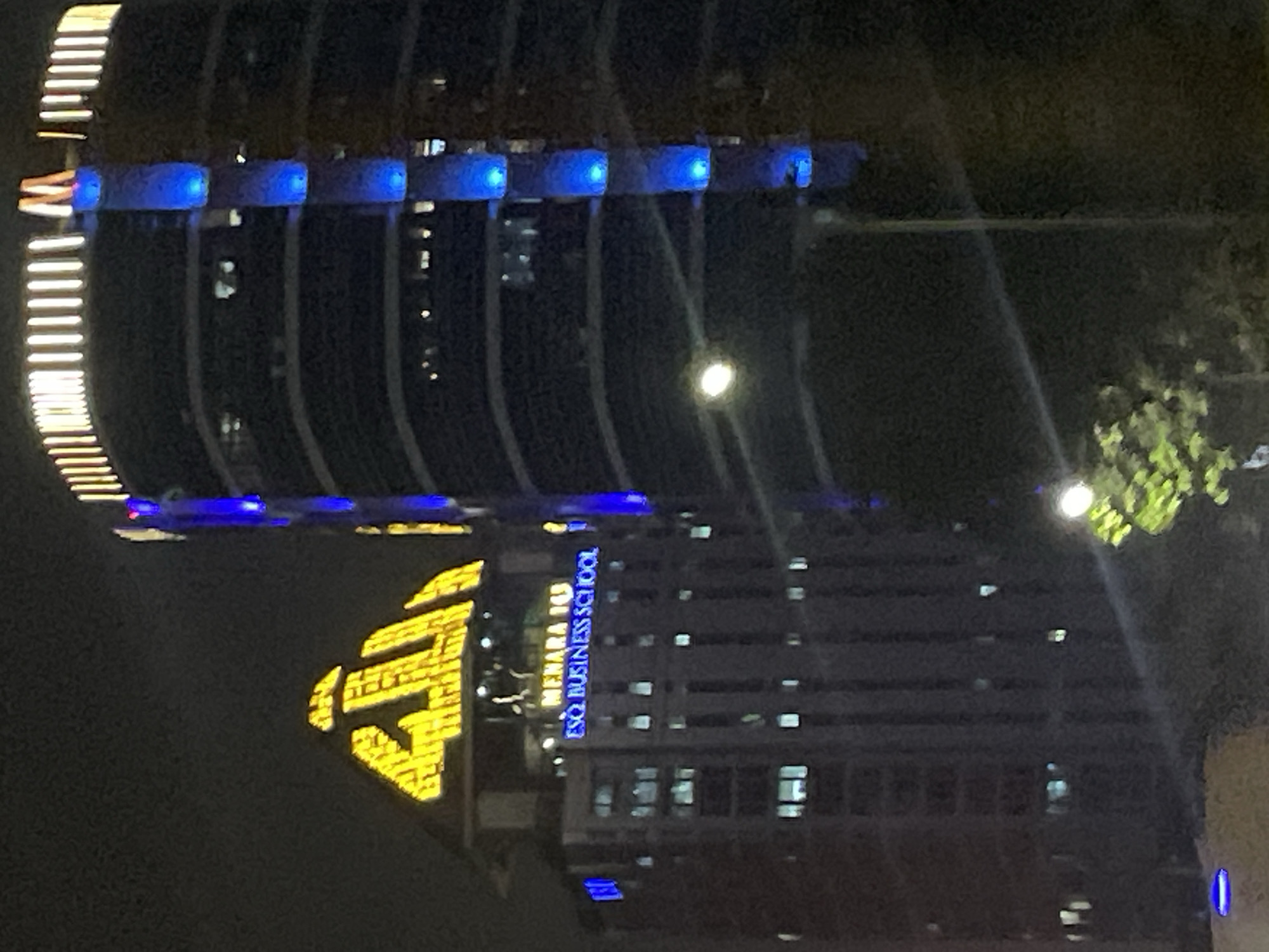 The image shows two prominent buildings at night in South Jakarta, Indonesia. The building on the left has a large, illuminated Arabic inscription in yellow on its upper section, appearing to be "Allah". Below this inscription, the building displays the text "MENARA 165" and "ESQ BUSINESS SCHOOL" in blue lights. The building on the right is a taller, modern structure with a curved facade. It features vertical blue lighting elements and a horizontal strip of white lights at its top, with an orange "M" shape also visible at the very top. Streetlights and the lights from within the buildings are also visible. A tree with green foliage is partially visible in the foreground. The overall scene depicts an urban environment at night, illuminated by architectural lighting.
