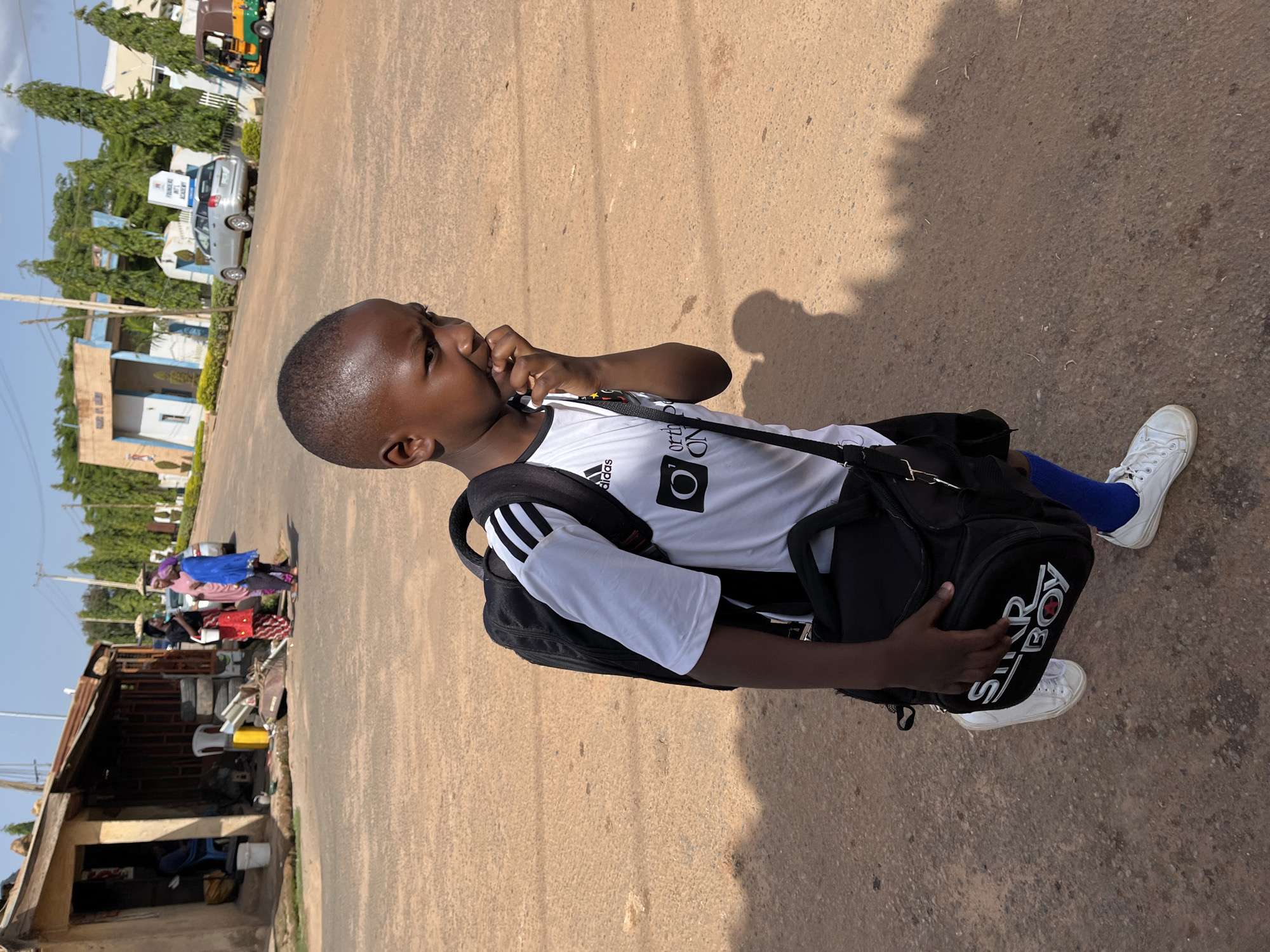 A young boy, wearing a white Adidas t-shirt with black stripes on the sleeves and black shorts, is walking outdoors. He has a black backpack slung over his shoulder and is holding his right hand up to his mouth, seemingly biting his nails. He is also wearing blue socks and white sneakers. The scene appears to be a street in Barakin Akawo, Nigeria, with a sandy, dusty road. In the background, there are buildings, trees, and a car, suggesting an urban or semi-urban environment. The sun is shining, casting shadows on the ground, indicating it is daytime. The boy's expression is neutral. The backpack has the text "STAR BOY" and a red cross symbol printed on it.