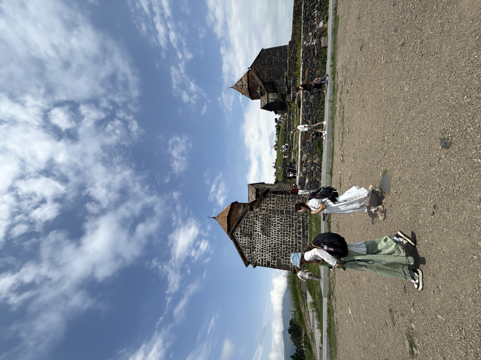 The image depicts the Sevanavank Monastery complex, situated in the city of Sevan, Armenia. In the foreground, several individuals are walking on a gravel path. A woman wearing a light blue hat, a white long-sleeved top, and a flowing green skirt is in the center-left, carrying a black backpack. To her right, another woman in a white dress with puffed sleeves and a black backpack walks away from the camera. Further back, other tourists can be seen near the stone structures of the monastery. The monastery itself, constructed from dark stone with a conical red roof and a cross on top, is visible in the middle ground. It is perched on a hill overlooking a body of water, likely Lake Sevan, although the water is not directly visible in this frame. The sky above is a clear blue with scattered white clouds, suggesting fair weather. The overall scene captures a moment of tourism and exploration at a historical and religious site.