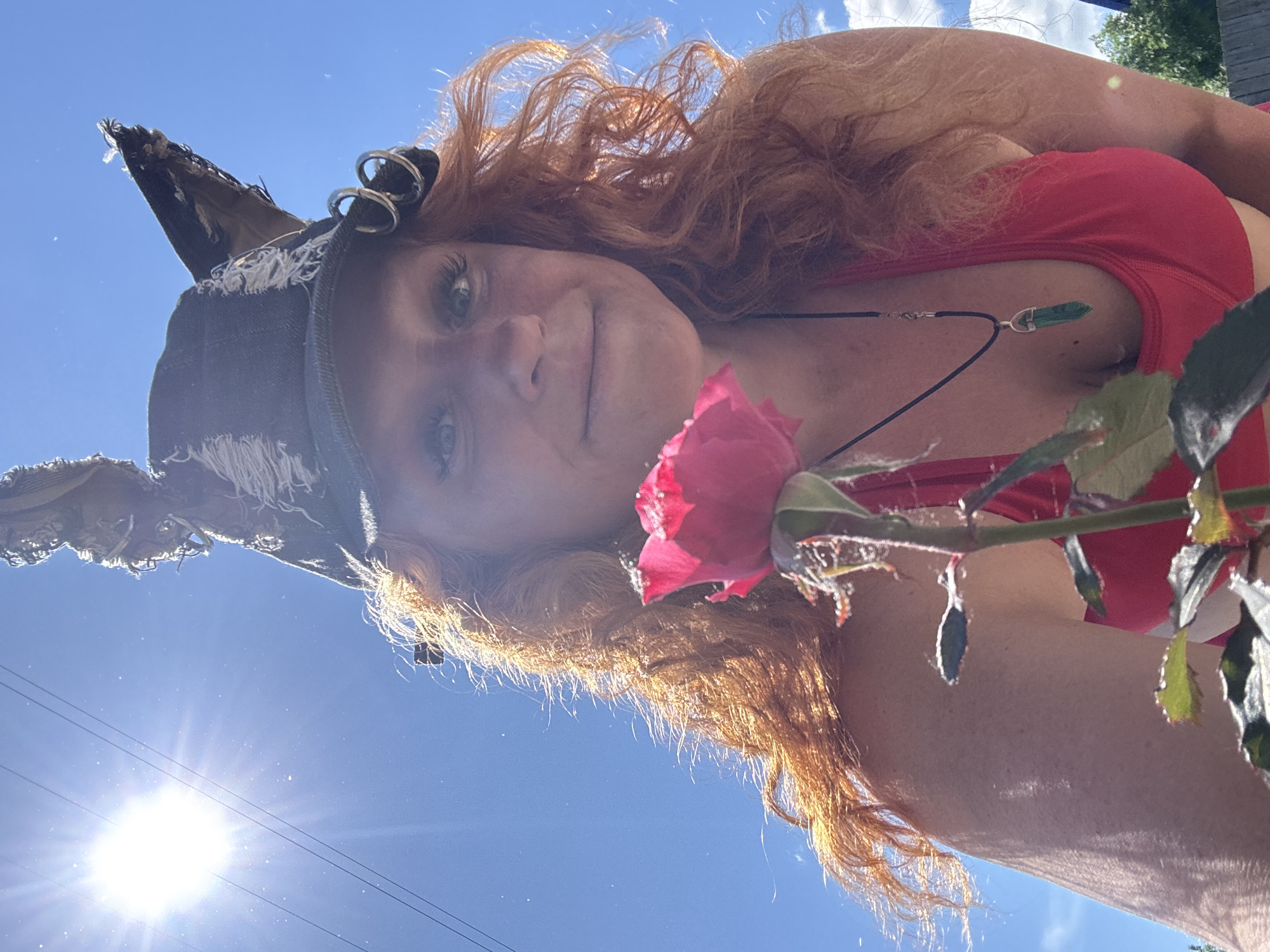 This media file shows a close-up, upside-down shot of a person with long, wavy, red hair and fair skin. They are wearing a red top and a distressed denim hat with grommets and frayed edges. A dark necklace with a green, crystalline pendant hangs around their neck. A single pink rose is held close to their face, with its petals slightly blurred. The background is a clear blue sky with bright sunlight, suggesting it is daytime. The sun's glare creates lens flare effects across the image. In the lower portion of the image, power lines are visible against the sky. The overall mood appears casual and perhaps a little playful, given the unusual angle and the presence of the flower and hat. The location is identified as Chaikovsky, Russia.