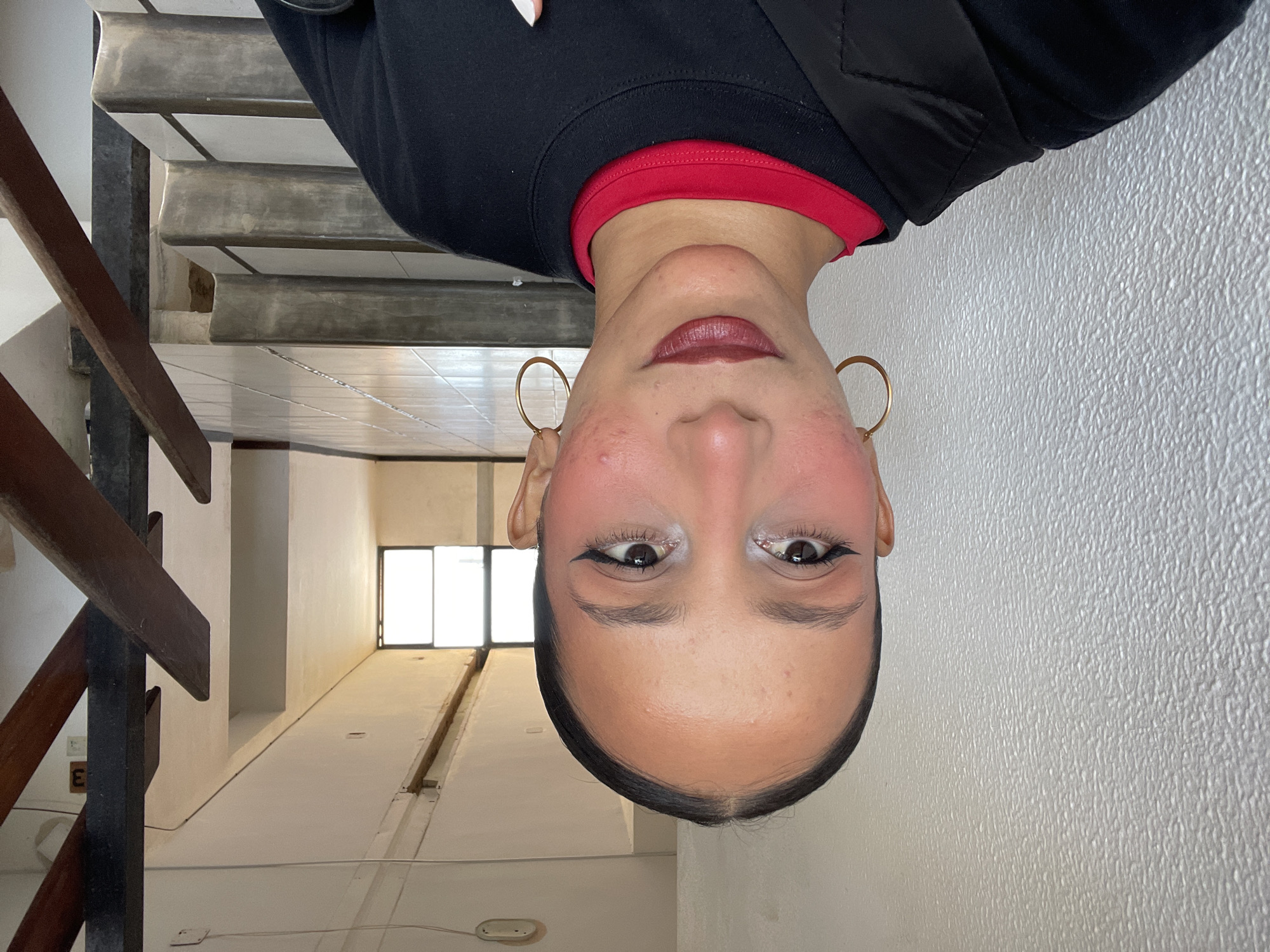 A medium shot captures a young woman facing the camera, positioned indoors against a textured white wall on the left. She has dark hair pulled back, a clear complexion with some minor blemishes, and is wearing makeup including winged eyeliner, blush, and dark lipstick. She sports large gold hoop earrings and a black sweatshirt layered over a red shirt. The background reveals a staircase with wooden railings and tiled steps, leading up to a hallway with windows. The environment appears to be a residential or commercial building, possibly an apartment complex. No specific indicators place the location in Porlamar, Venezuela.