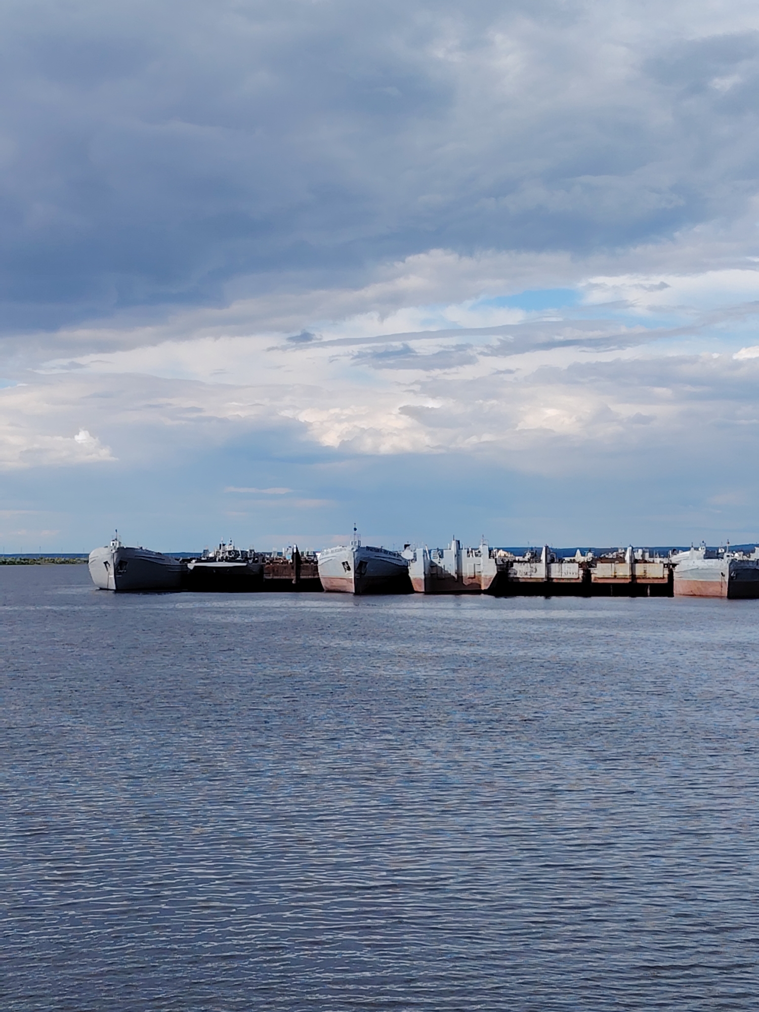 The image shows a line of interconnected, inactive ships or barges docked in the water near Chaikovsky, Russia. The water is choppy with small waves, and the sky is filled with a mix of dark and light clouds. In the distance, a faint green line could indicate land. There are no people visible in the image.