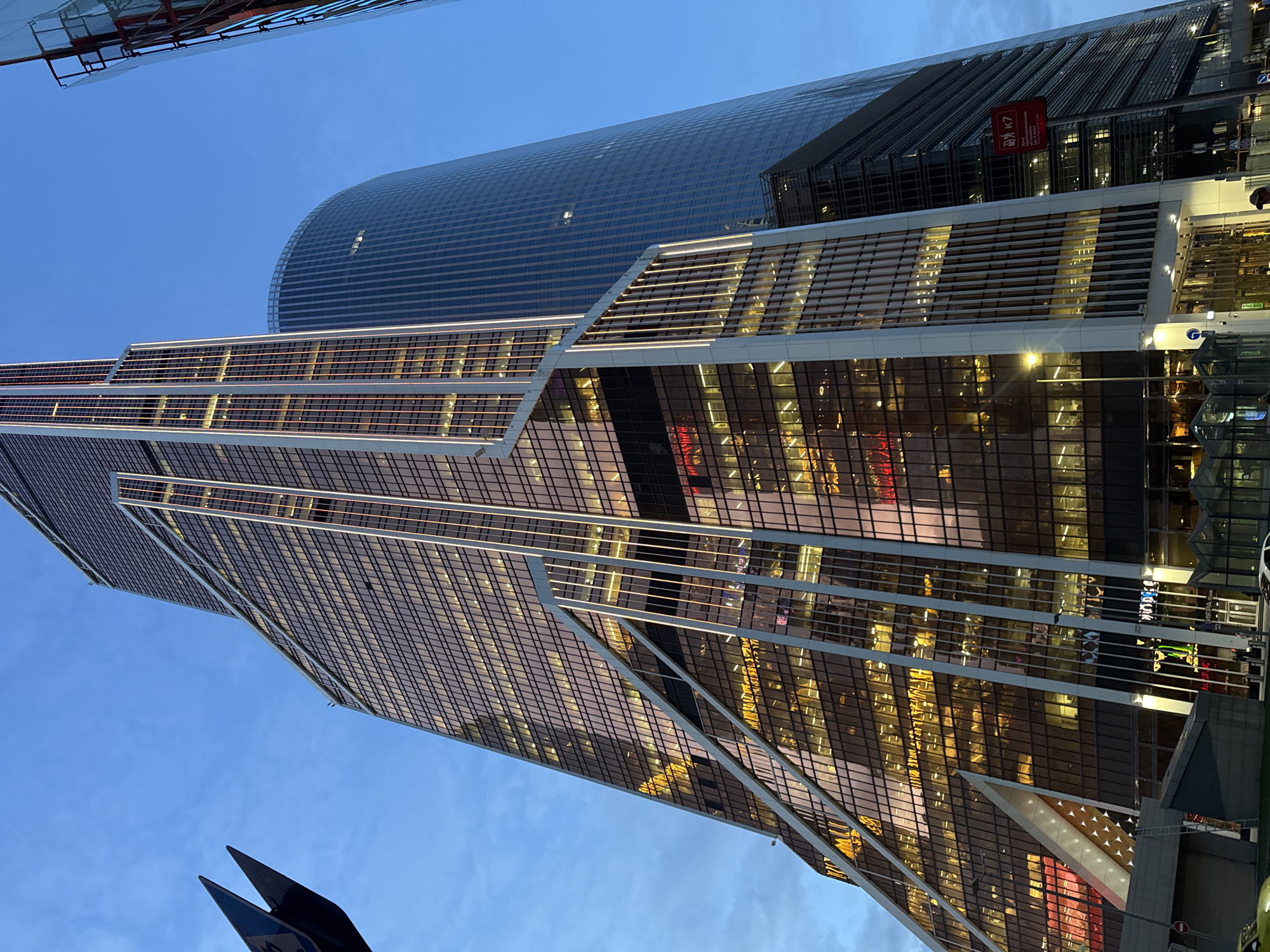 The image captures a striking urban landscape at dusk or early night, dominated by two modern, glass-clad skyscrapers against a clear deep blue sky. The primary building in the foreground is a complex, multi-tiered structure featuring a facade of numerous reflective glass panels that glow golden from interior lighting. White LED strips accentuate its architectural lines and angles. Behind it, a tall, curved, dark-blue glass building rises, its smooth surface reflecting the sky.
No explicit human activity is depicted, but the illuminated windows suggest active commercial or office spaces. The overall scene conveys a sense of contemporary urban development.
Notable details include:
* The dramatic contrast between the illuminated golden reflections of the main building and the dark, reflective surface of the curved building.
* The intricate patterns formed by the glass panels and the linear white lights.
* In the bottom-left foreground, abstract black angular shapes are visible, possibly part of a sign or sculpture.
Visible text on a prominent red sign attached to the main building includes:
* "K7"
* A pictogram of a person.
* Text below that appears to be in Russian, reading "30 этажей" (30 floors), "Рестораны" (Restaurants), "Кафе" (Cafes), "Магазины" (Shops), and "Тренажерный зал" (Gym).
Another sign on the lower right part of the building shows partially readable text, possibly "O'CLOCK" and other blurred letters.
The clear sky and the interplay of natural and artificial light suggest calm weather conditions. Given the architectural style and the Russian text, the location context of Khimki, Russia, is consistent with the scene.