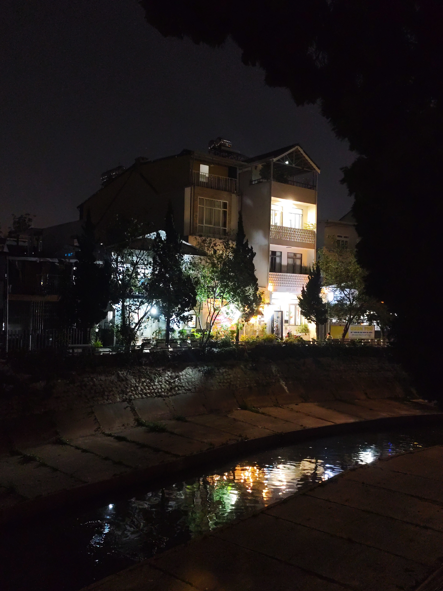 At night, a three-story building is seen illuminated on the opposite side of a concrete drainage canal filled with water reflecting the lights. Trees and shrubbery line the embankment, partly obscuring the ground floor of the building. The sky is dark. The location is Da Lat, Vietnam. No people are visible in the image.