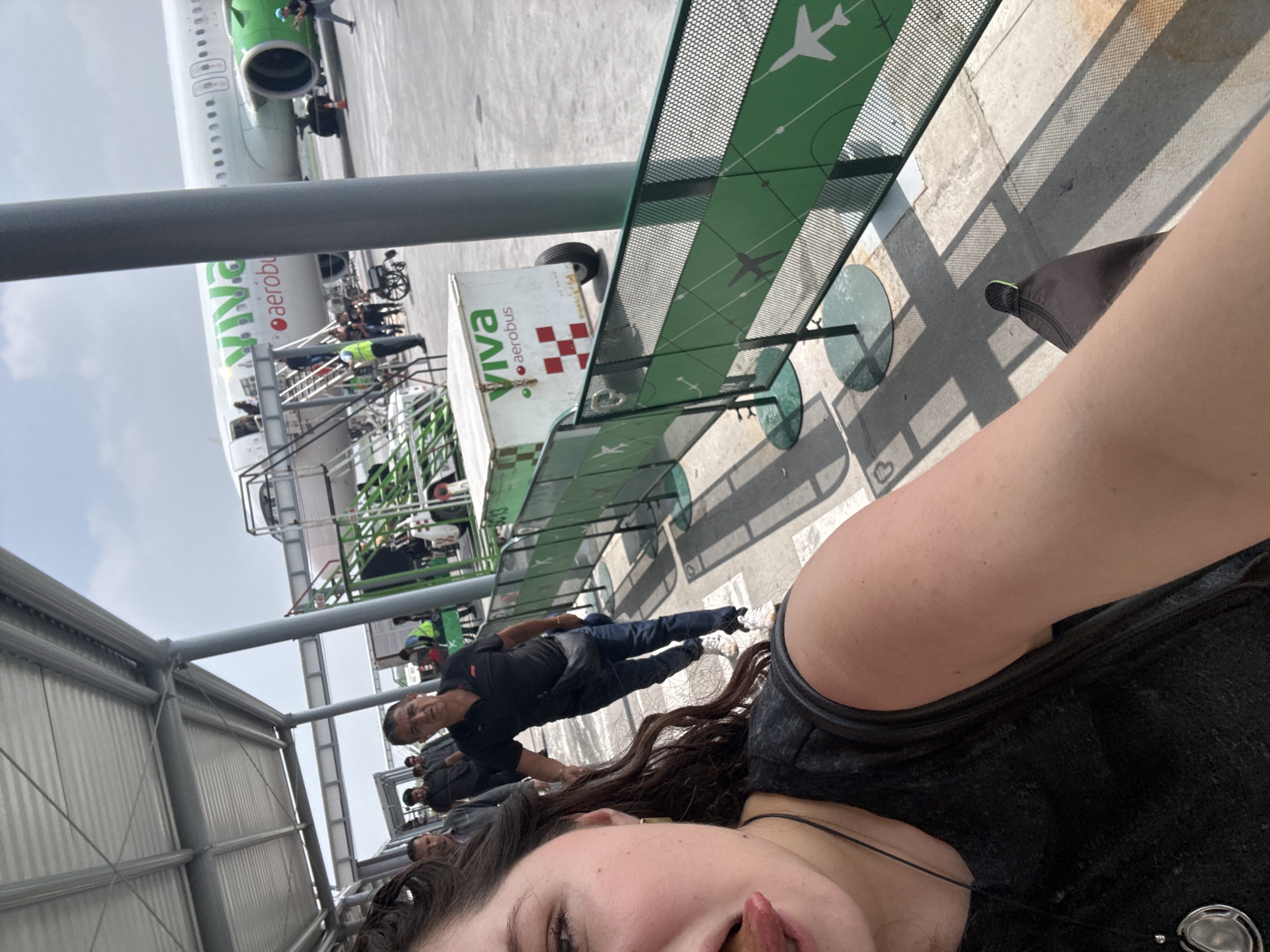 A rotated, eye-level outdoor shot shows a woman in the foreground, possibly taking a selfie. Her face, curly hair, black shirt and jewelry are partially visible. Behind her are other people, including a man in a black shirt looking towards the camera. In the background, a Viva Aerobus airplane sits on the tarmac, with passengers boarding via stairs. The boarding process and other airport personnel are visible. A Viva Aerobus branded luggage cart is also nearby. A green metal fence with airplane images runs along a walkway. The location appears to be an airport in Apodaca, Mexico, identified by the Viva Aerobus branding.