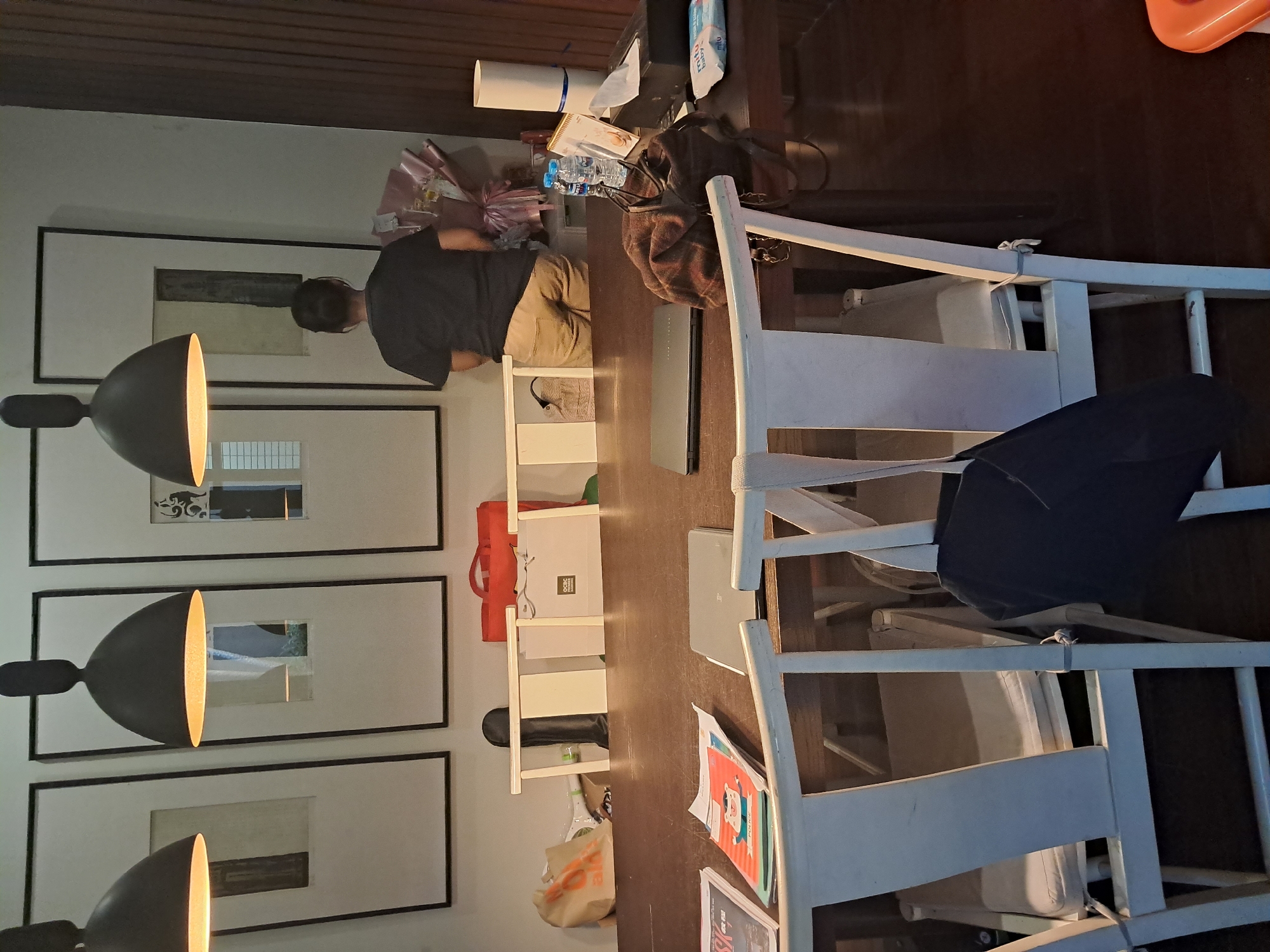 The image captures an indoor scene, likely in Bareng Lor, Indonesia, focusing on a dining table and its surroundings. A person, viewed from the back, stands near a wall adorned with three large framed artworks and hanging pendant lamps. The individual wears a dark t-shirt and tan pants and appears to be interacting with a bouquet of flowers.
The table is laden with various items, including two closed laptops, a patterned cloth bag, a tissue box with a dispenser, a stack of papers, and two bottles of water. In the foreground, two white chairs with cushioned seats are partially visible. One chair has a dark bag hanging from its back.
Behind the table, on a shelf or partition, several objects are placed, including a red tote bag, a light-colored shopping bag, and a white cylindrical object. The overall environment suggests a domestic or semi-professional setting with a mix of personal and functional items. The lighting appears to be a combination of artificial light from the lamps and ambient room light.
