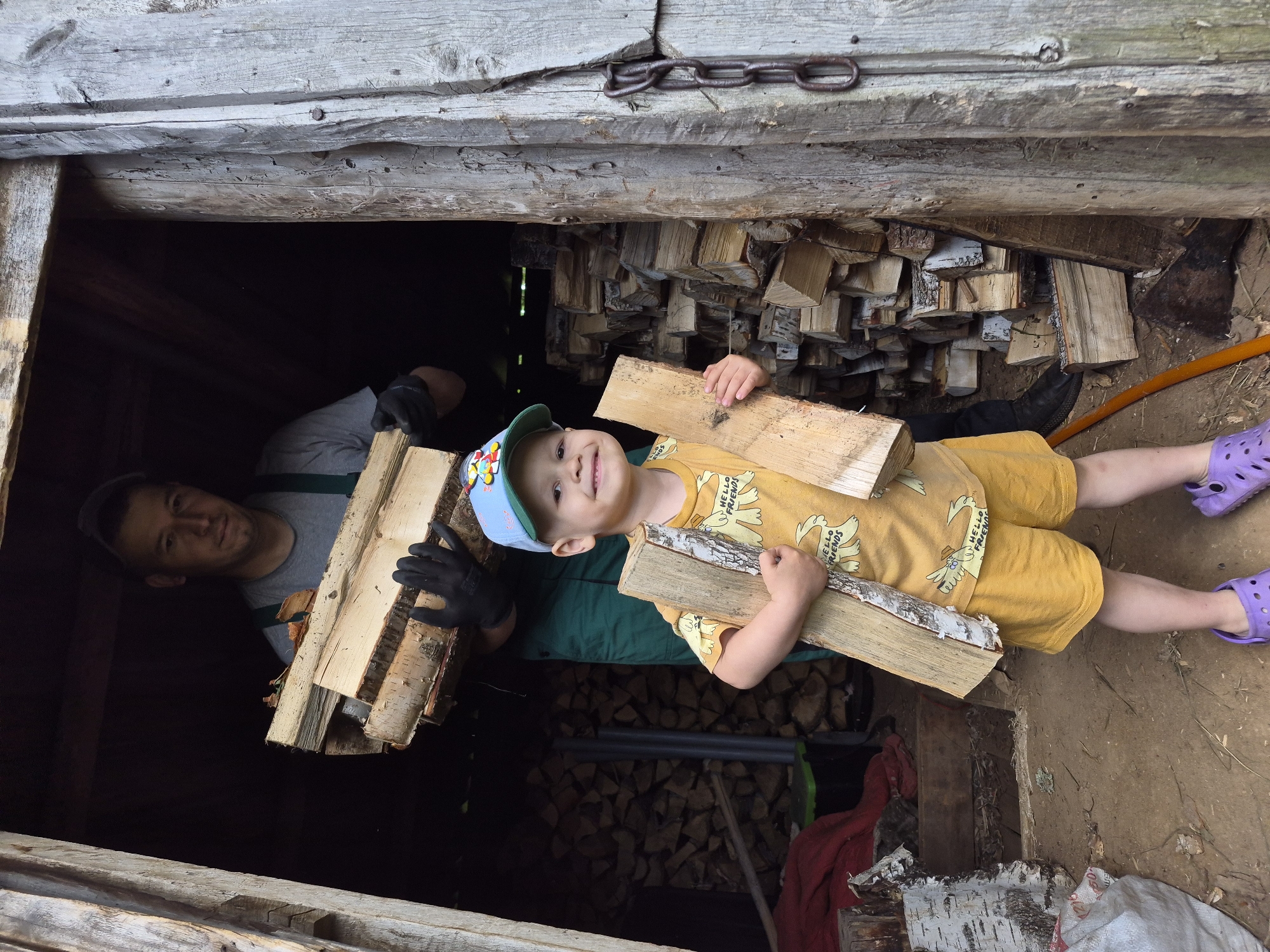 In a rustic, wooden structure filled with stacked firewood, a man and a young boy are engaged in the task of wood gathering. The man, wearing a grey t-shirt and green overalls, stands in the background, carrying a substantial bundle of chopped wood. He is wearing black gloves, suggesting he is actively involved in the labor. In the foreground, a young boy, sporting a yellow t-shirt with a cartoon print and yellow shorts, proudly holds two pieces of firewood. He wears a blue baseball cap and purple clogs. The environment appears to be outdoors, with a dirt floor and the weathered exterior of the wooden building. The presence of chains on the right side of the structure hints at its functionality, possibly as a storage shed or barn. The overall scene depicts a shared chore between father and son in a rural setting. The location is identified as Kharnavy, Russia.