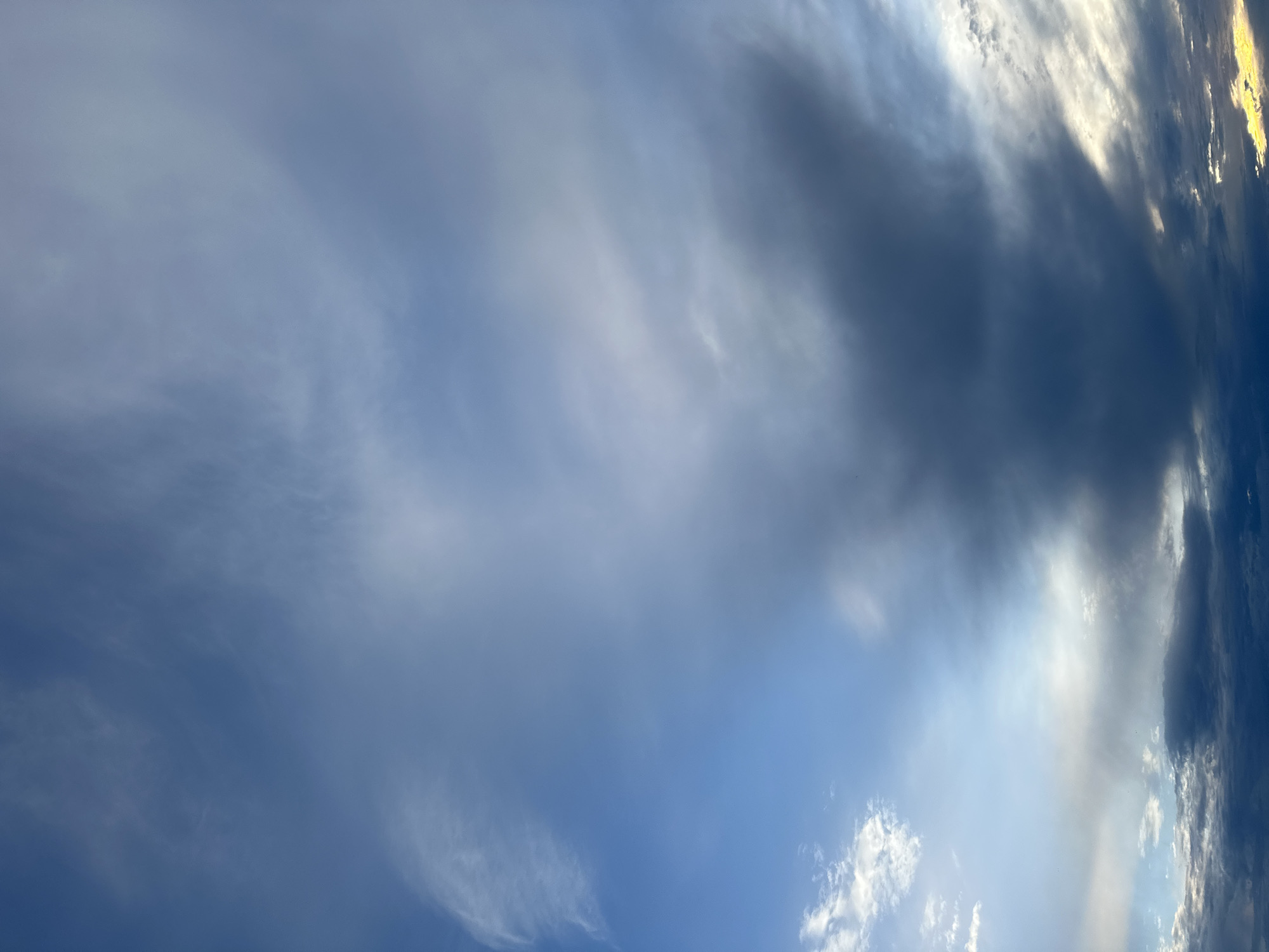 The image shows a sky with varying cloud formations. On the left, there are thin, wispy clouds spread across a blue sky. Towards the right, the clouds become denser and darker, appearing gray and shadowing part of the sky. There are also small patches of white, puffy clouds in the lower right corner. A bright, hazy light seems to shine through the clouds, possibly the sun. No other subjects, objects, or actions are visible in the image. The location is not discernable.