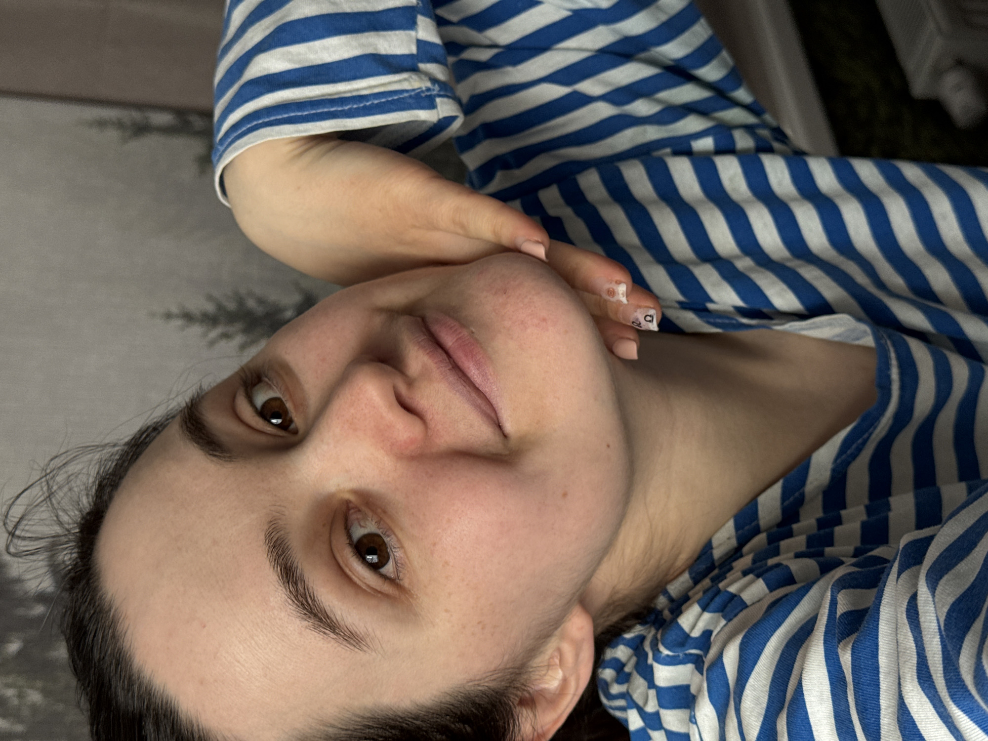 A close-up portrait of a person, likely female, is presented, rotated ninety degrees clockwise, giving the impression of the subject lying down or looking upwards. The person has dark brown hair, which appears to be pulled back, framing a face with light skin, brown eyes, and natural eyebrows. Small, scattered freckles are visible across the nose and cheeks. Her lips are a natural pink hue, slightly parted. Her expression is neutral, calm, and direct towards the viewer.
She is wearing a top with prominent horizontal blue and white stripes. Her left hand is gently resting against her jawline and cheek, with the fingers slightly curled. Her fingernails are painted a pale pink or nude color. On one of the fingernails, small, stylized black letters or symbols are visible, appearing to be 'D' and 'U'.
The background is softly blurred and appears to be an indoor setting, showing a greyish, textured surface with faint, dark, abstract patterns, possibly a rug or blanket. The overall atmosphere conveyed is relaxed and natural. This media file was captured in Izhevsk, Russia.