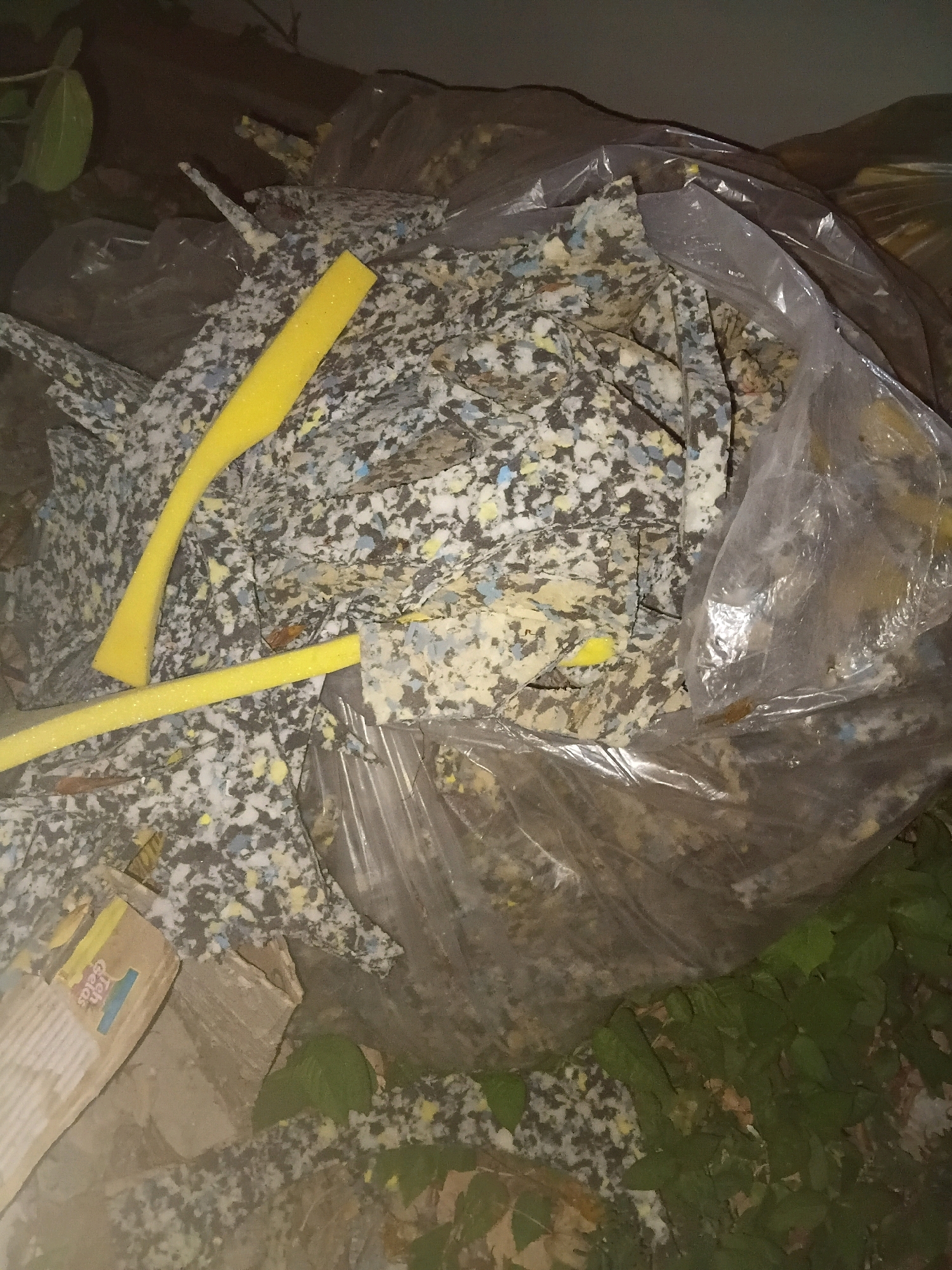 The image depicts a pile of discarded foam material, appearing to be insulation or packing foam, contained within a clear plastic bag. The foam is a mixture of various colors, including gray, white, yellow, and blue speckles. Two long, yellow foam tubes are visible protruding from the pile. Some cardboard pieces are also present on the left side of the image, with partial text visible on one piece, appearing to be "Teh Gelas". The scene appears to be outdoors, with some green foliage visible in the lower right and upper left corners, suggesting it is a natural or semi-natural environment. The lighting is dim, indicating it may be nighttime or a shaded area. The location is identified as Cikal, Indonesia.