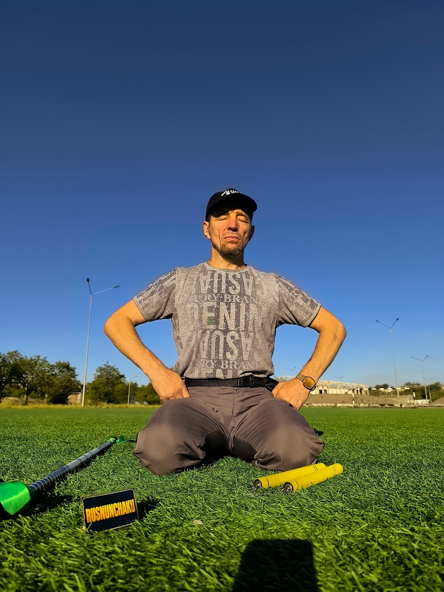A man wearing a black baseball cap and a grey t-shirt with a graphic print is kneeling on a bright green artificial turf field. He has his hands on his hips and his eyes are closed. To his left is a black and green nunchaku, and to his right are two yellow nunchaku. A small rectangular sign with the text "RUSNUNCHAKU" in yellow lettering on a black background is placed in front of the black and green nunchaku. The background shows a clear blue sky, some trees, and distant stadium structures with streetlights. The scene appears to be outdoors in a city park or sports complex.