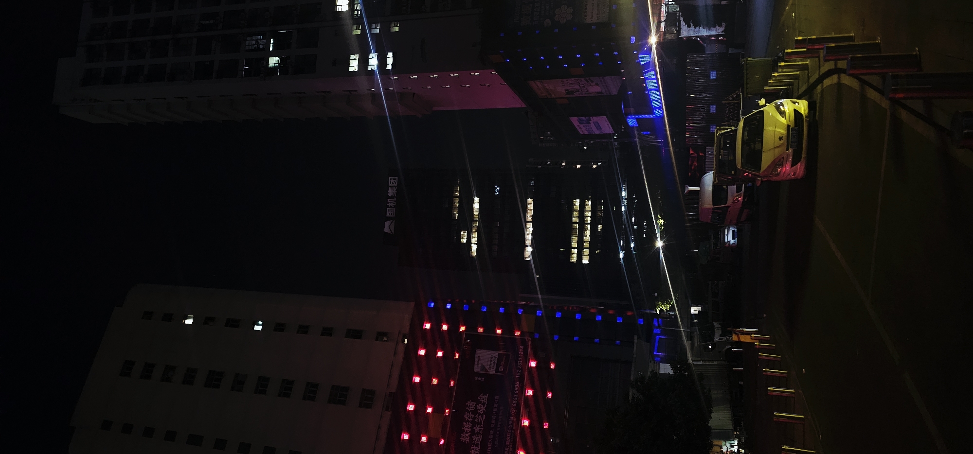 The image is an angled, nighttime shot of a city scene in Jiulongpo District, China. The foreground features a large, multi-story building with its windows illuminated. Below this building, a street is visible with several vehicles, including a yellow and white car that appears to be a taxi or service vehicle, and another white vehicle behind it. Red and blue lights are decorating the facade of another building in the mid-ground, possibly indicating an advertisement or signage. The background shows more illuminated buildings and a dark sky, suggesting a typical urban environment at night. The overall impression is one of a busy, lit-up city. Visible text on a poster in the mid-ground reads: "数据存储 就选东芝硬盘" which translates to "Data Storage Choose Toshiba Hard Drive." Further text includes contact information and company names in Chinese.