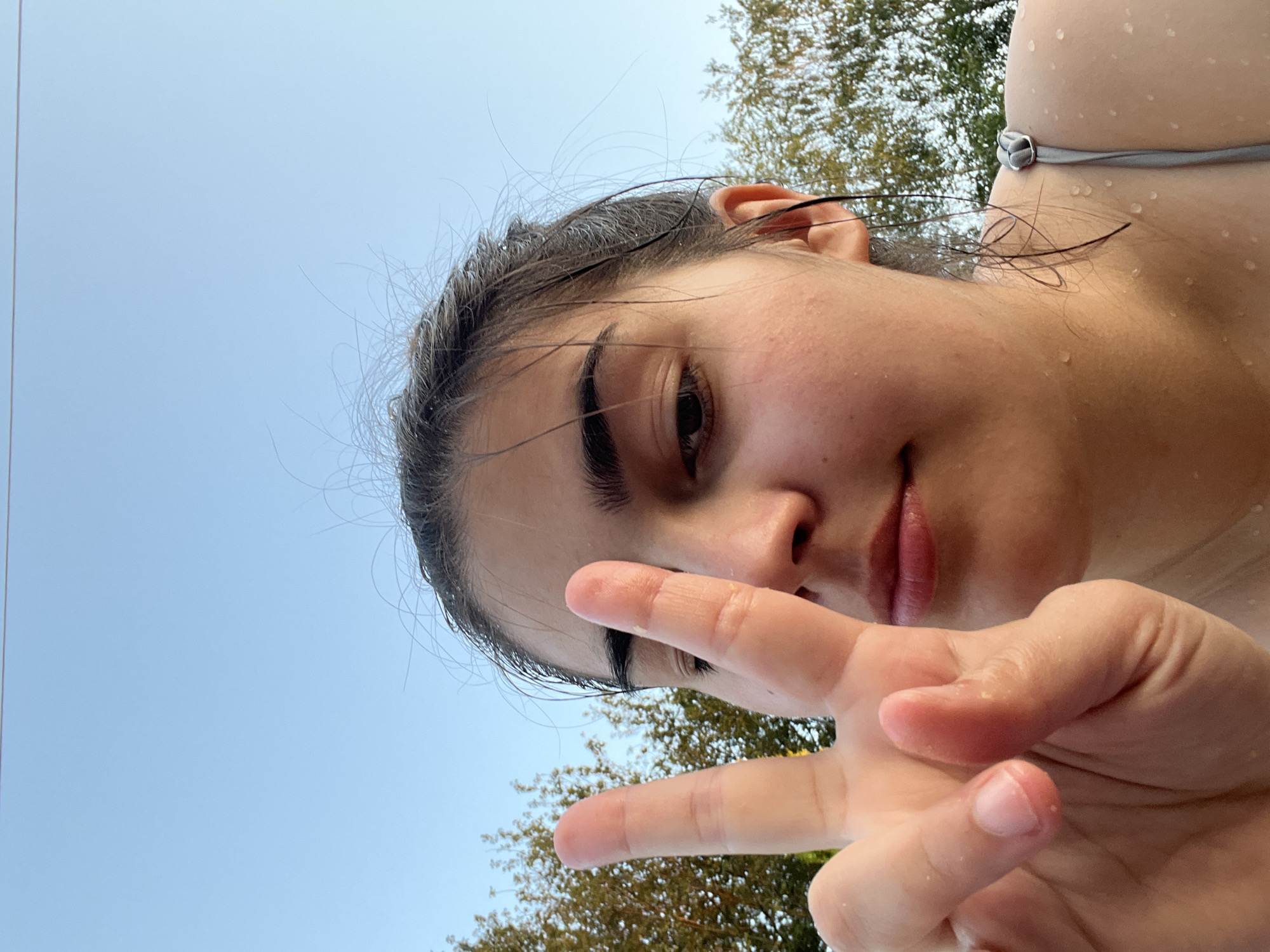 A close-up, low-angle selfie shows a young woman with wet, dark hair and clear skin. She is outdoors with a bright blue sky in the background and trees on either side. She holds up her left hand, forming a peace sign with her index and middle fingers extended. Water droplets are visible on her skin and hair, suggesting she may have been swimming. She is wearing a grey tank top with a strap visible over her shoulder. The scene appears to be in an outdoor recreational area.