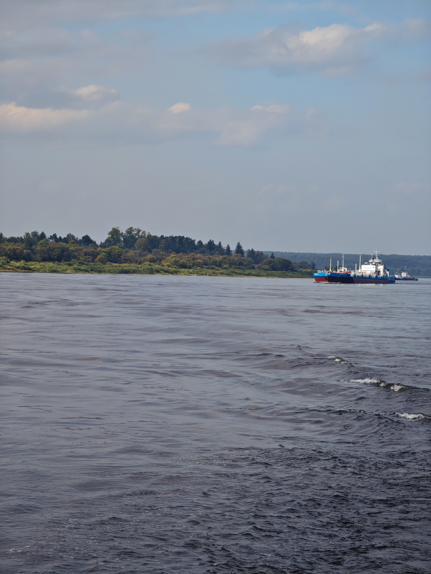 A wide river fills the foreground and midground of the image, its surface rippled by wind and the wake of a passing barge. The barge, a blue and red vessel with a white superstructure, travels away from the viewer on the right side of the river. It is followed by a smaller tugboat. On the far bank of the river, a dense line of trees forms a natural border, with a few scattered houses visible amongst the foliage. The sky above is a pale blue, dotted with scattered white clouds. The overall scene depicts a tranquil day on a Russian river, likely in the city of Verkhne-Pashino, given the context.
