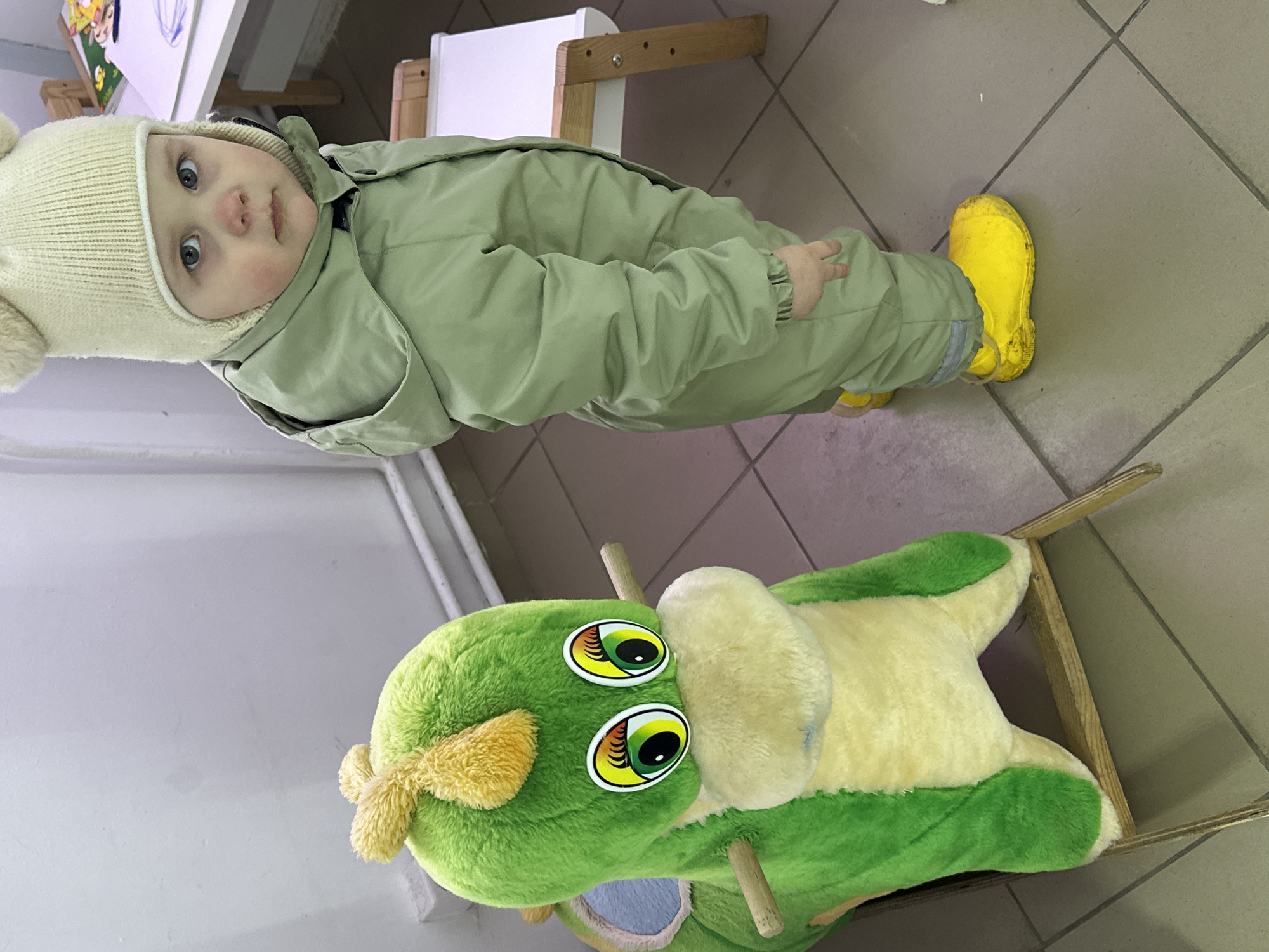 A baby wearing a cream-colored hat and light green jumpsuit with yellow boots stands next to a plush rocking horse toy in a room. The rocking horse is green and yellow. The floor is tiled and there is a white table with wooden legs nearby. The location is likely Chaikovsky, Russia.