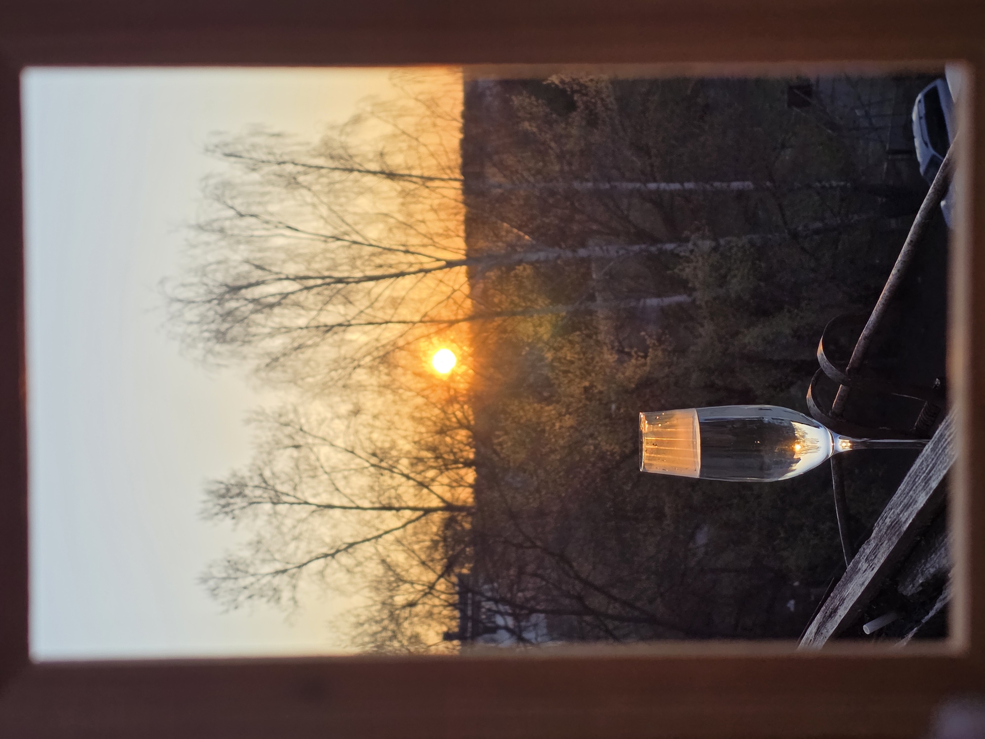 A glass of clear liquid rests horizontally on a wooden railing, likely of a balcony, framed by a wooden window frame. Outside the window, a sunset is visible, with a bright orange sun positioned near the center of the frame, partially obscured by bare tree branches. The sky transitions from a pale orange near the horizon to a lighter color higher up. On the right side, the trees are darker, silhouetted against the fading light. The location is likely Chaikovsky, Russia, based on the presence of birch trees and architecture. A dark-colored vehicle is vaguely visible behind the trees.