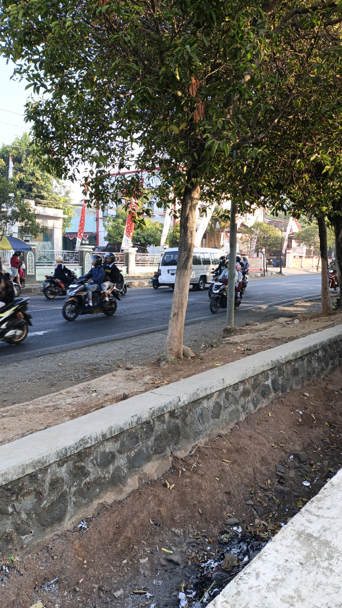 In the city of Kuwasen, Indonesia, a paved road runs through a residential area lined with trees and buildings. The scene is captured from a slightly elevated perspective, looking across the road and towards the buildings. A stone and concrete retaining wall runs along the edge of the road, with a dry, debris-filled ditch below. Several motorcycles, ridden by people wearing helmets, are traveling on the road. A white van is also visible, parked on the side of the road. The trees in the foreground and background are lush with green leaves, with some reddish-brown leaves also present. The sky is clear and bright, suggesting it is daytime.