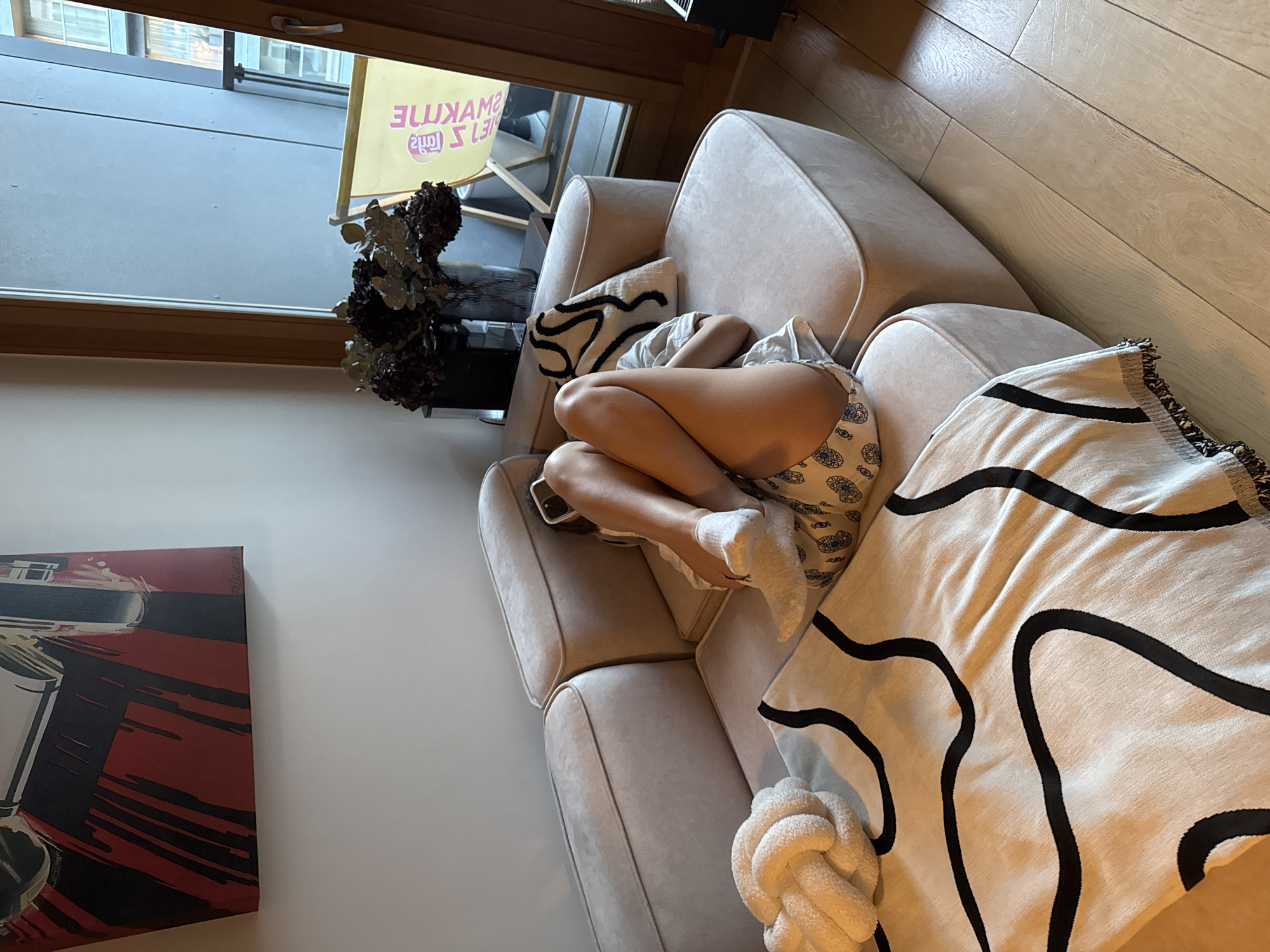 A person is seen resting or sleeping, curled up on a light beige sofa in what appears to be a modern living space. The individual, likely female, is wearing a light-colored top, patterned shorts with a distinctive eye-like motif, and white socks. Their body is comfortably positioned with knees drawn up, conveying a sense of relaxation.
The sofa is upholstered in a soft, light-toned fabric, featuring clean lines and plush cushions. A cream-colored blanket with bold, black, wavy abstract patterns is draped over a section of the sofa. Next to the person's feet, a white, intricately knotted decorative pillow adds to the cozy aesthetic. To the left of the sofa, near a large window, sits a dark vase containing dried, dark-colored flowers or branches.
Through the window, which has a light wooden frame, an outdoor scene is visible, suggesting a balcony, patio, or an adjacent building. The natural light filtering in indicates it is daytime. A light wooden floor is visible on the right side of the image. On the far left wall, a segment of an abstract painting in red and black hues can be seen.
A notable detail in the background is a yellow banner or sign visible through the window, possibly part of an outdoor piece of furniture or an advertisement. The text on this banner appears mirrored or reversed, reading "SELJ (2MUA) CAMELUX" from right to left as seen. The overall scene depicts a peaceful and comfortable indoor environment.