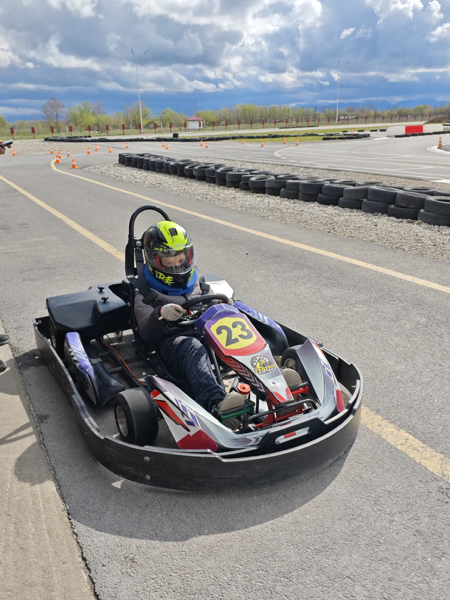 The image shows a child in a go-kart on an outdoor track in what appears to be Kishpek, Russia, based on the Cyrillic text on the go-kart. The child is wearing a helmet and appears ready to drive. The track is surrounded by tires and orange cones. A building, trees, and cloudy skies are visible in the background. The kart has the number 23 on it. The track is asphalt.