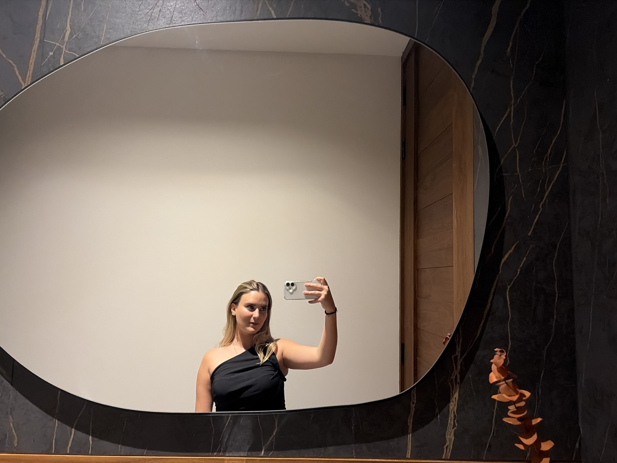 A young woman with light blond hair and a black one-shoulder top is taking a selfie in a mirror. The mirror is irregularly shaped with a black marble-like frame. Behind the woman, reflected in the mirror, is a beige wall and a wooden door. The right side of the mirror's frame is surrounded by a black marble wall with light brown veins. A decorative plant stem with orange discs is placed on the counter to the right of the mirror. The location is San Bernardino Tlaxcalancingo, Mexico.