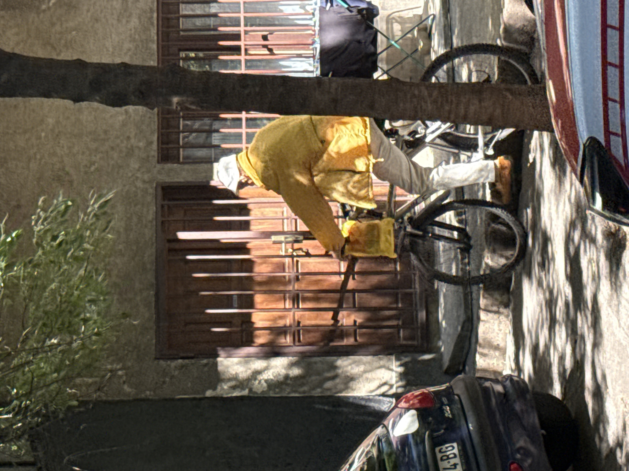 A man wearing a white cap and a yellow jacket rides a bicycle past a building in Belgrade, Serbia. The bicycle has a yellow bag attached to the front. The man is wearing light-colored pants and brown shoes. The building has a wooden door with vertical slats and metal bars. A dark-colored car is parked to the left of the frame. The ground is paved and has dappled sunlight and shadows. A tree trunk is positioned to the right of the man and bicycle.