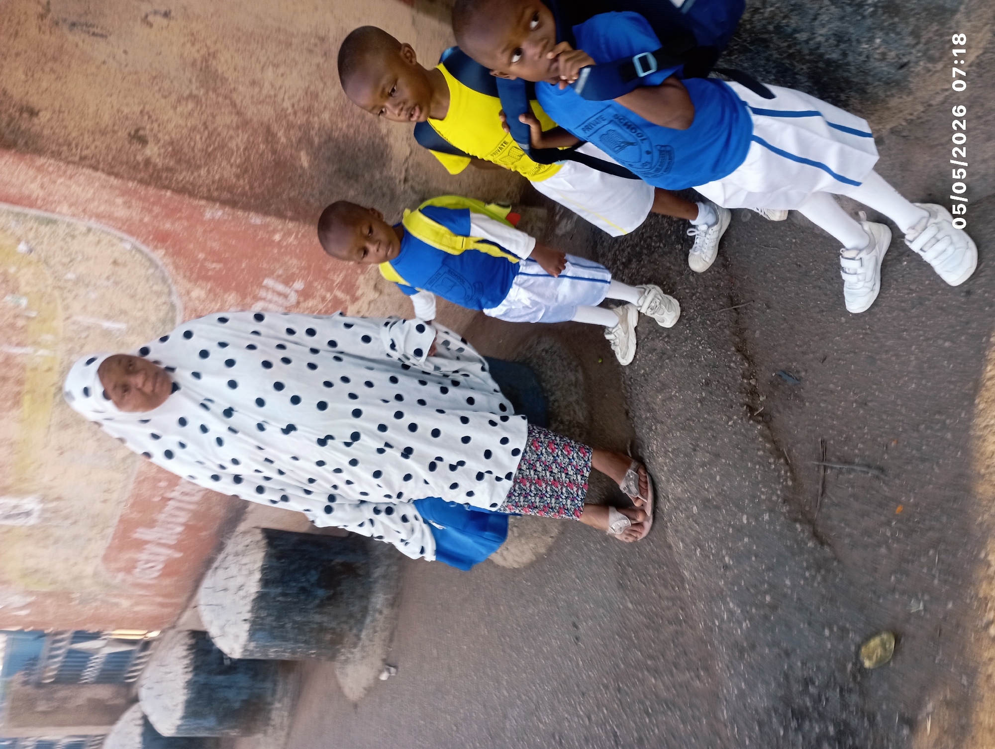The image shows a woman and three children standing on a paved surface. The woman is wearing a white headscarf and a white gown with black polka dots. The children are wearing school uniforms that are a combination of blue, yellow, and white. Two of the children have blue tops with the words "Private School" on them. All three children have white sneakers. The background appears to be a building facade with faded colors. The image was captured on 05/05/2026 at 07:18. The location is likely the city of Jos, Nigeria.