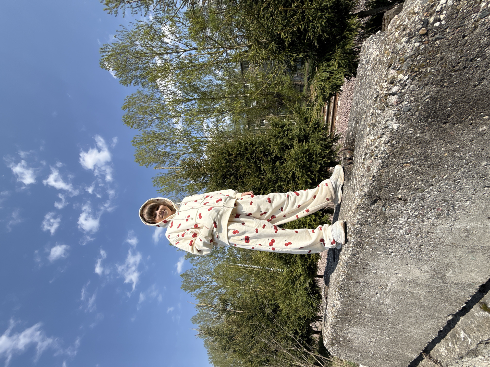 A person is standing on a large, gray concrete structure in what appears to be an outdoor environment. The person is wearing a cream-colored, hooded tracksuit adorned with red cherry patterns. Behind the structure are trees and a building visible in the background under a blue sky with scattered clouds. The scene is potentially in Shebunino, Russia, but without further information, this can’t be proven.