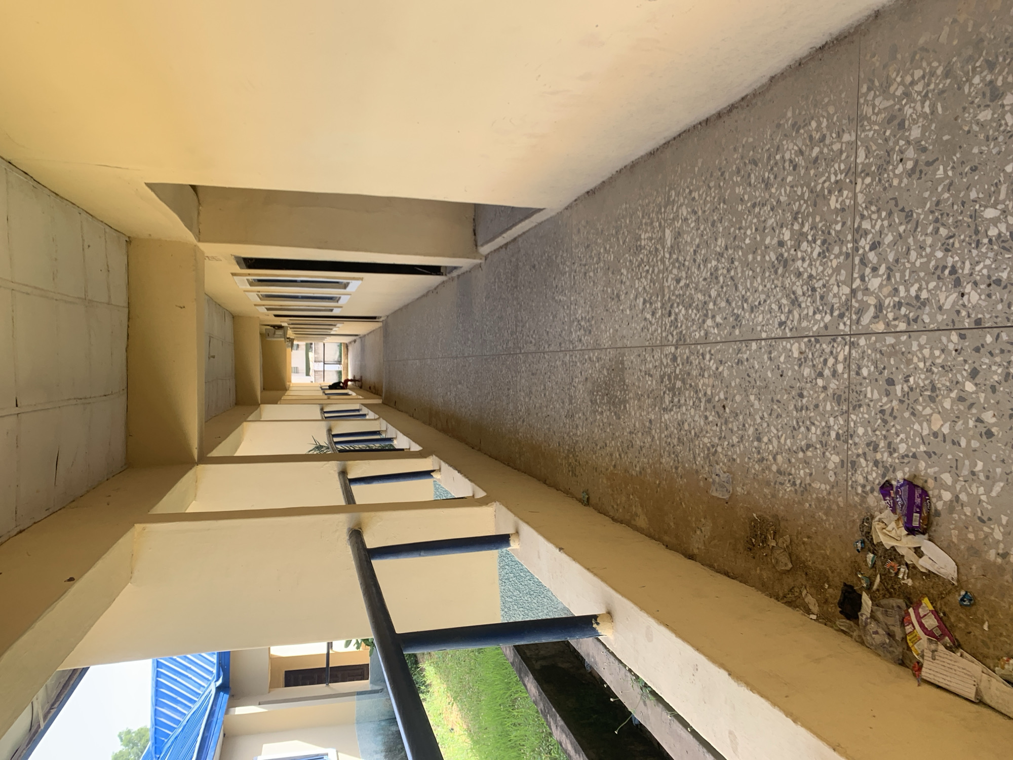 The image depicts an outdoor hallway or walkway with a yellow-painted building on the right and a row of windows on the left, leading to a distant opening. The walkway has a speckled, terrazzo-like floor and is lined with a yellow railing supported by blue posts. To the left of the railing, there is a strip of green grass and a concrete drain. Debris, including crumpled wrappers and paper, is scattered on the ground near the railing. The ceiling of the walkway appears to be made of suspended panels. In the distance, a person is seated on a bench, and another building with a blue roof is visible. The overall impression is of an institutional or commercial building. The location is identified as Jalingo, Nigeria.