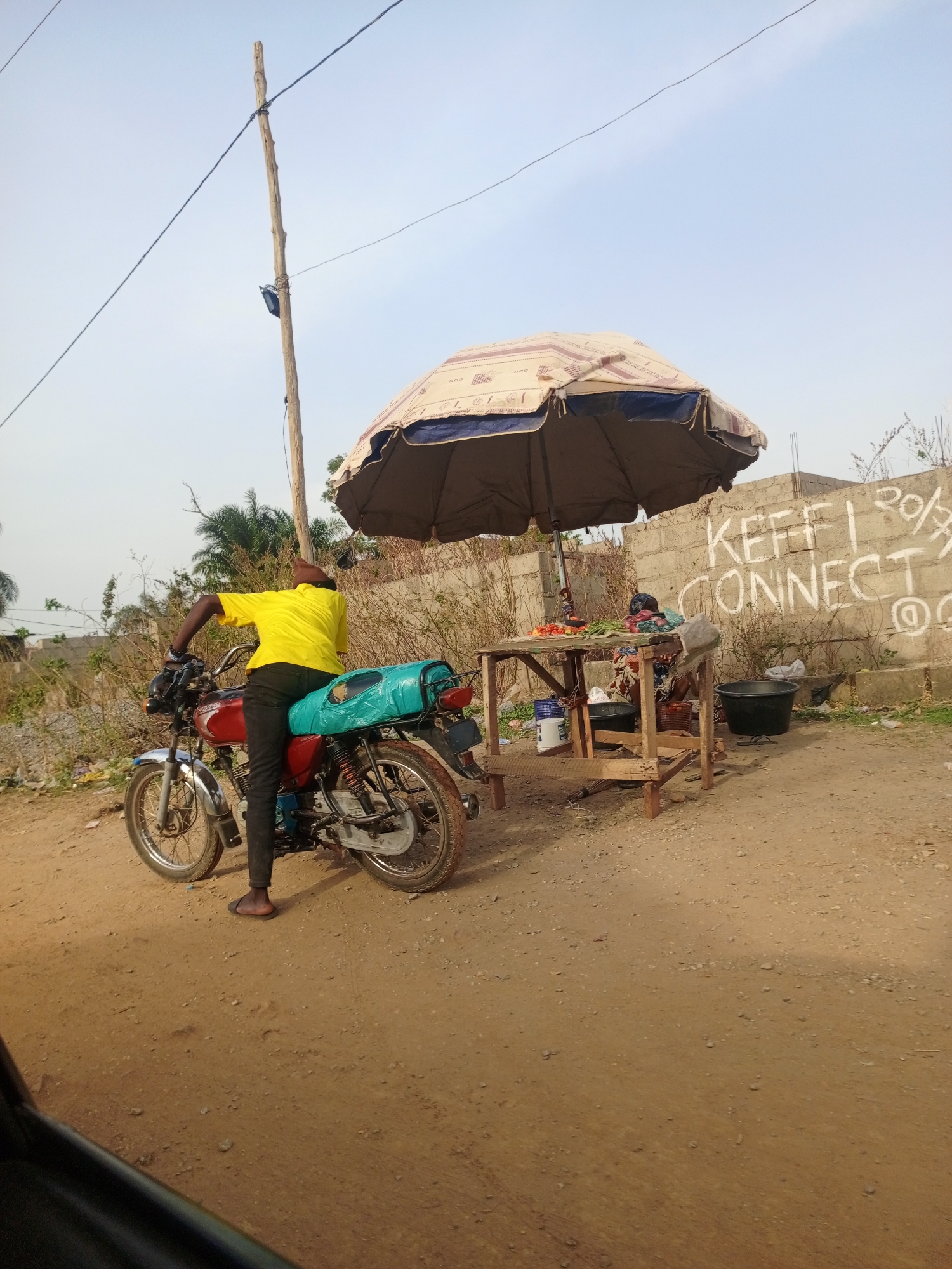 A medium-shot displays a man on a motorcycle purchasing goods from a roadside vendor in Unknown, Unknown. The man, wearing a bright yellow shirt and dark pants, sits on a red motorcycle with a turquoise bundle strapped to the back seat. He is positioned on a dirt road facing the vendor. The vendor's stall is shaded by a large, slightly tattered umbrella, and the stall is arranged with various goods, including tomatoes and green vegetables. A woman, likely the vendor, sits behind the stall. Behind the stall is a concrete wall with "KEFFI CONNECT" spray-painted on it. A wooden pole with electrical wires attached stands adjacent to the motorcycle. The sky is clear, and the environment suggests a rural or developing area.
