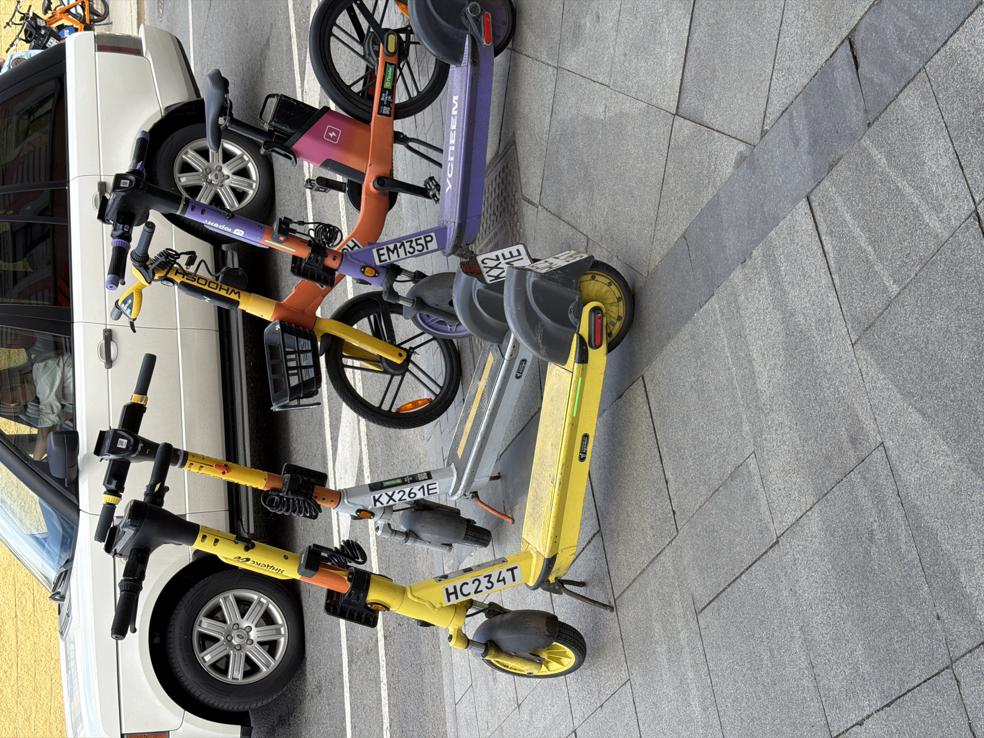 The image shows a row of shared electric scooters and bicycles parked on a paved surface next to a white vehicle. The scooters and bikes are arranged in a line, with some leaning against the vehicle. The scooters are various colors including yellow, grey, and purple. The bicycles are orange. License plates are visible on the scooters. The location appears to be an urban area in Moscow, Russia.