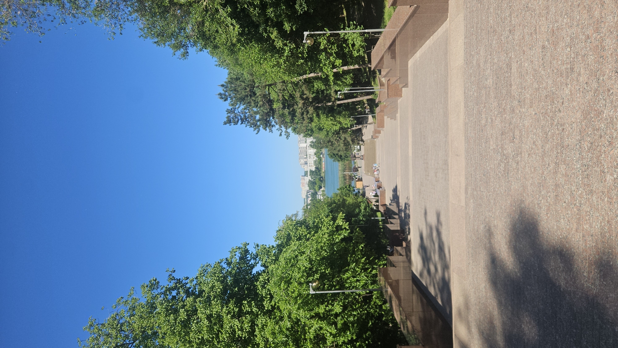 The image shows a view overlooking a city lake in Chișinău, Moldova, taken from an elevated position. The foreground features a wide, lightly colored stone walkway or plaza with steps leading down towards the lake. A low wall runs along the right side of the walkway. Green trees flank both sides of the walkway, partially obscuring the view. Street lamps are positioned among the trees. In the distance, across the lake, are several multistory buildings. The sky is clear and blue. People are visible near the lake shore.