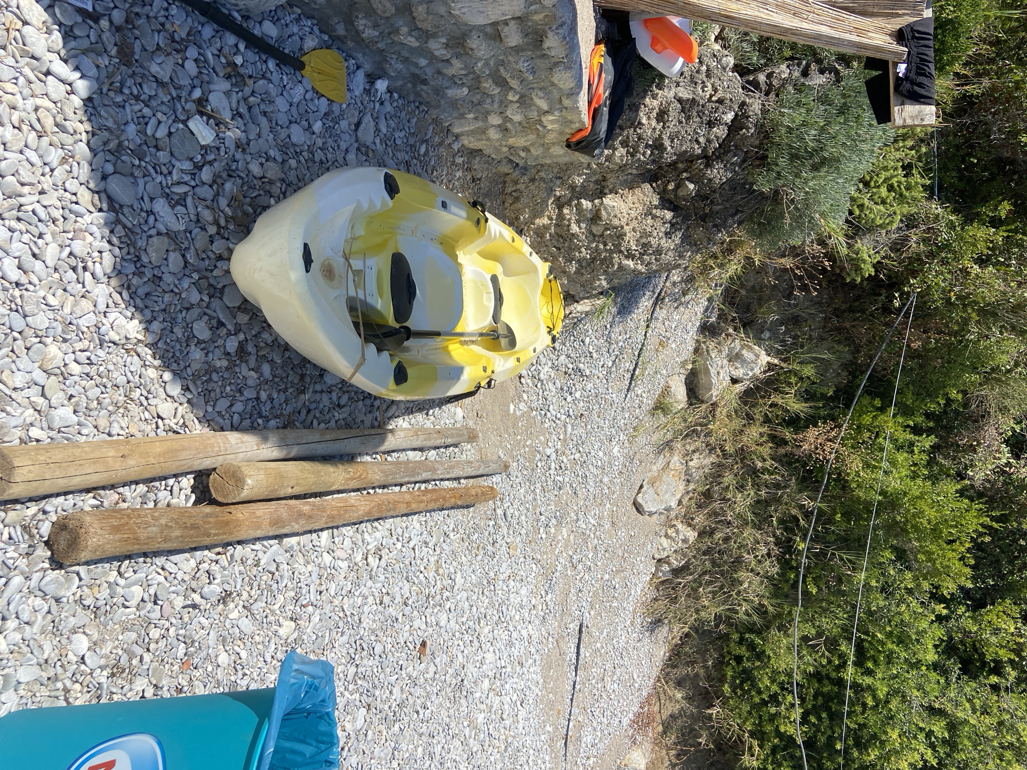 The image depicts a bright, outdoor scene, likely a beach or rocky shoreline, with lush green vegetation covering an incline in the background. In the foreground, a yellow and white kayak rests on a bed of smooth, grey pebbles. A black and yellow paddle lies next to the kayak. To the left, a low stone wall is visible, with a bamboo structure leaning against it. A piece of black fabric is draped over a wooden beam attached to the bamboo. Orange and white buoys are situated on the stone wall. To the right of the kayak, three thick wooden poles stand upright in the pebbles. Further to the right, a bright teal trash can with a blue liner is partially visible. The scene is bathed in sunlight, casting shadows on the pebbles. The location is identified as Prijevor, Montenegro. There are no people visible in the image.