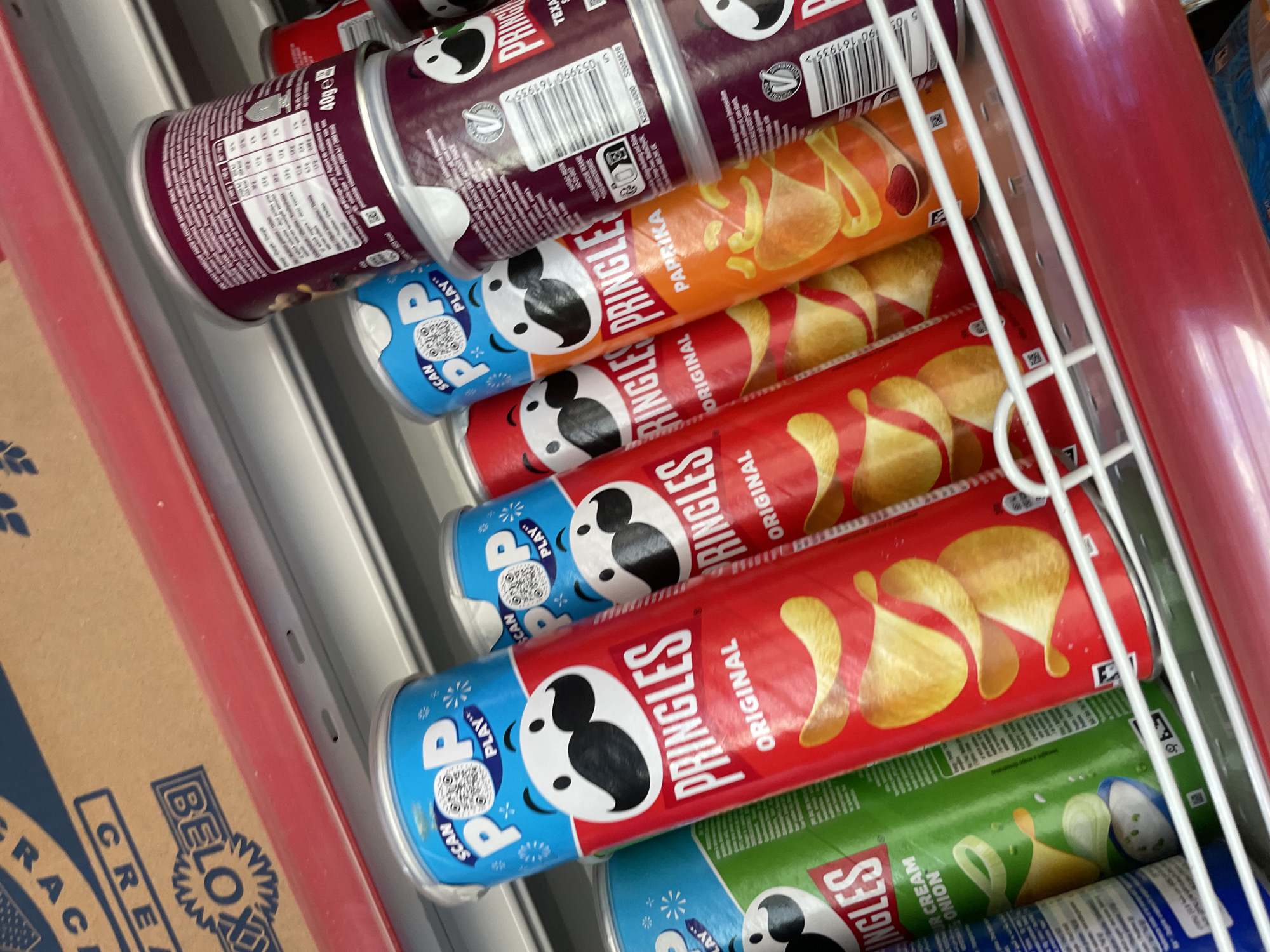 The image shows a close-up of a shelf filled with cans of Pringles potato chips. The cans are stacked on top of each other at an angle. The flavors visible include Original, Paprika, Texas BBQ Sauce, and Sour Cream & Onion. The cans have blue "scan POP play" labels on top.
There is a box with the text BELOX and CRE on the left. A part of a white metal shelf is visible between the cans. A red frame is visible on the left and right.
The location and people are not visible.