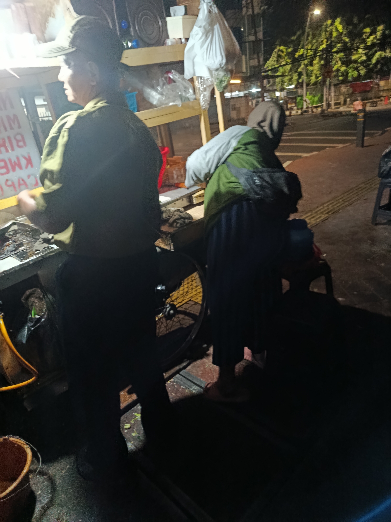 A medium shot captures a street food vendor preparing food at night in Central Jakarta, Indonesia. The vendor, wearing a dark cap and olive green jacket, is positioned in front of a cart filled with food items. To the right, a person wearing a green jacket and a headscarf is seated on a chair. The scene is illuminated by streetlights and the bright lights from the food cart, casting long shadows across the sidewalk and street. The background reveals a busy street with passing traffic, buildings, and lush green trees, indicating an urban environment. The overall atmosphere is one of a typical bustling night market in a Southeast Asian city.