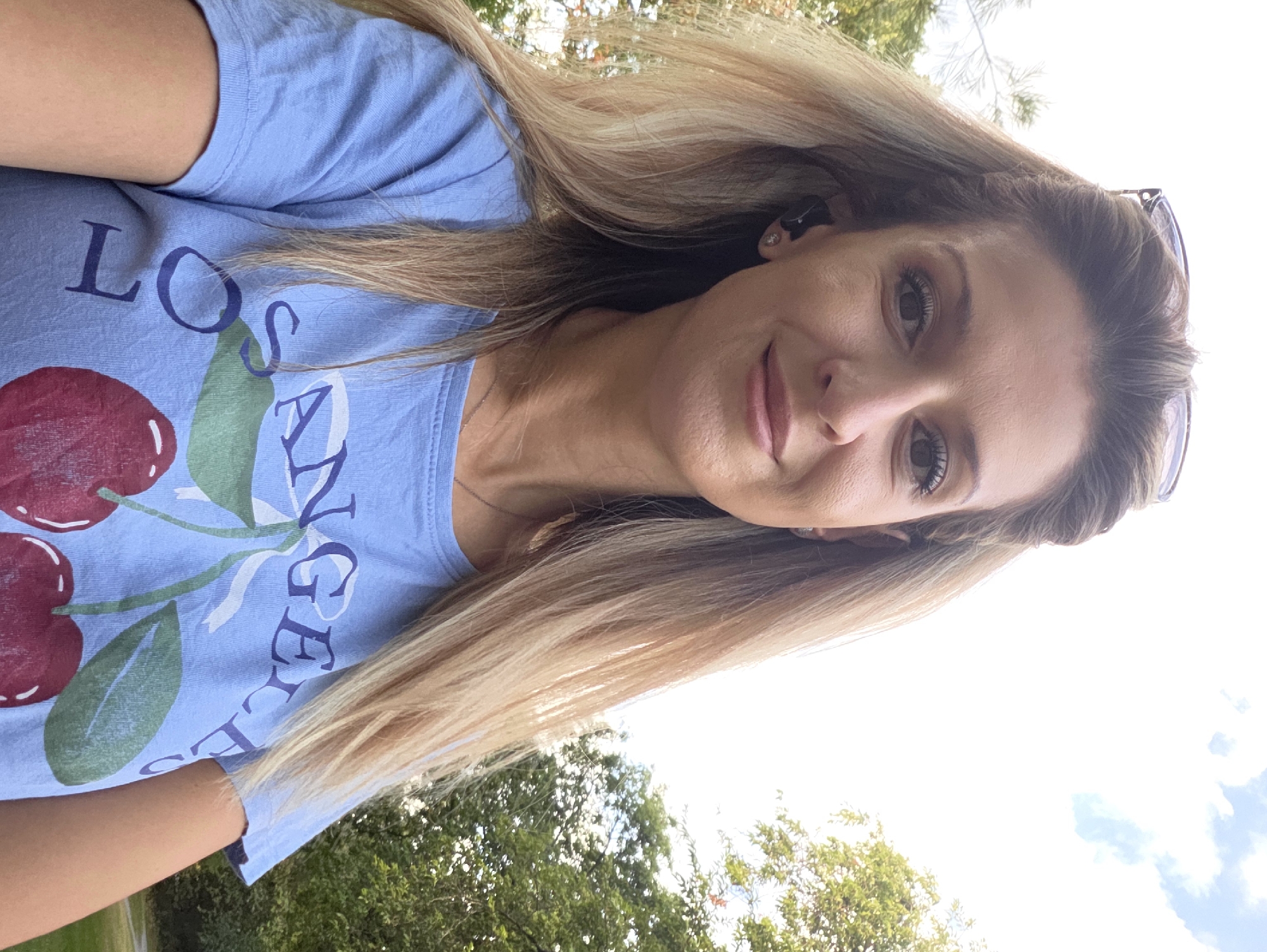 A woman with blonde hair pulled back, wearing sunglasses on her head and a light blue t-shirt with a cherry graphic, is taking a selfie outdoors. The background consists of trees and a bright sky with clouds, suggesting a park or a natural area. The image is taken from a slightly upward angle. There is no indication of the city of Munich, Germany.