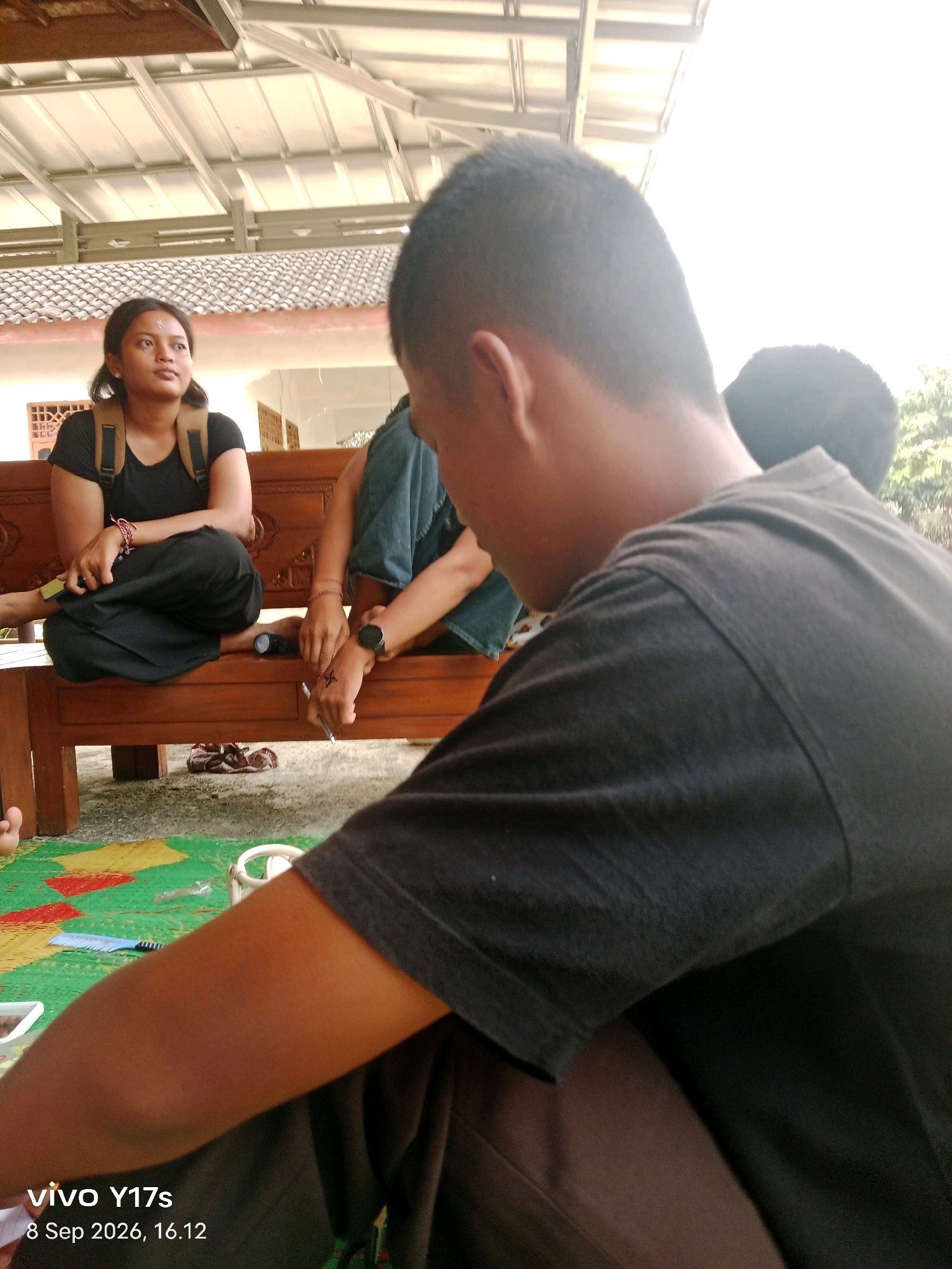 In Rama Dewa, Indonesia, a group of individuals are gathered outdoors, seated on a wooden bench. The scene appears to be in a semi-rural or residential area, with a building roof and some vegetation visible in the background. A young woman with dark hair, wearing a black t-shirt and dark pants, is seated on the left side of the bench, looking upwards and to her left. She has a backpack on. To her right, on the same bench, is a person wearing blue jeans and a dark t-shirt, with their legs crossed and a watch on their left wrist. This person is holding a pen and appears to be drawing or writing on their hand, which has a visible "X" marking. In the foreground, the back of a person wearing a dark t-shirt is visible, facing away from the camera and towards the others. Another person's leg is partially visible in the lower left corner of the frame. The ground is covered with a colorful mat. The overall impression is a casual social gathering.