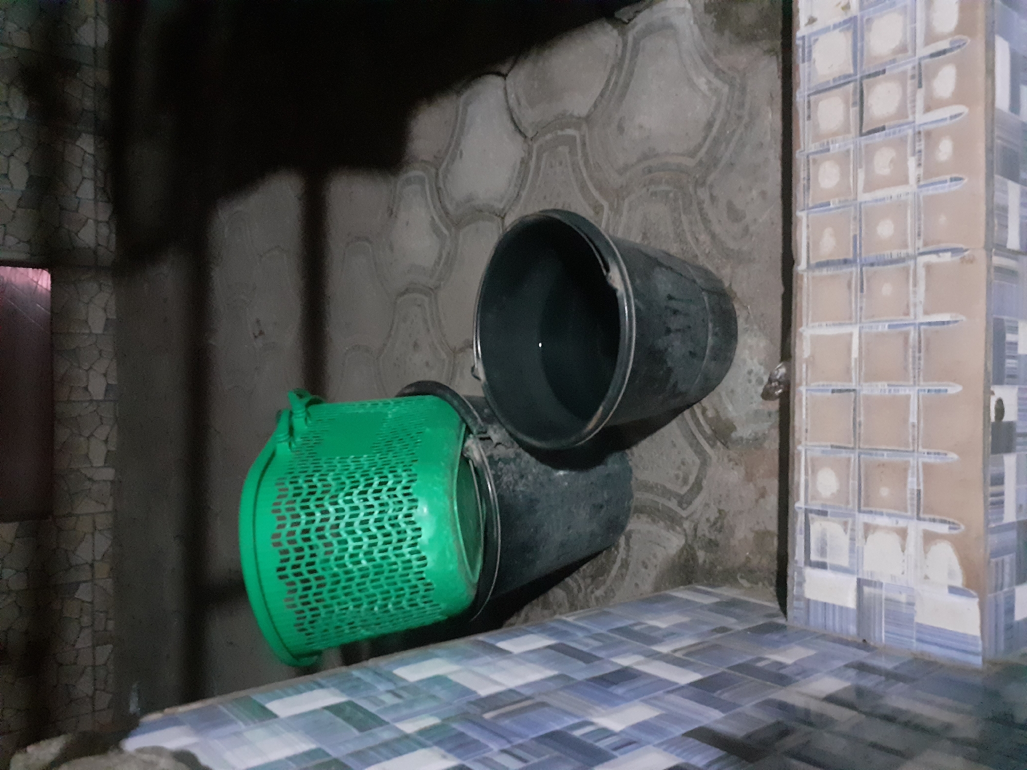 In Jalingo, Nigeria, two dark grey buckets and a green plastic basket are positioned on an outdoor tiled floor. One bucket, filled with water, sits to the right, while another, empty, is partially obscured by a green basket stacked on top of it. The surrounding area features a patterned tiled walkway in the foreground, with larger, interlocking grey tiles on the ground behind the buckets. A textured stone wall forms the backdrop, and the scene is illuminated by an artificial light source casting shadows.