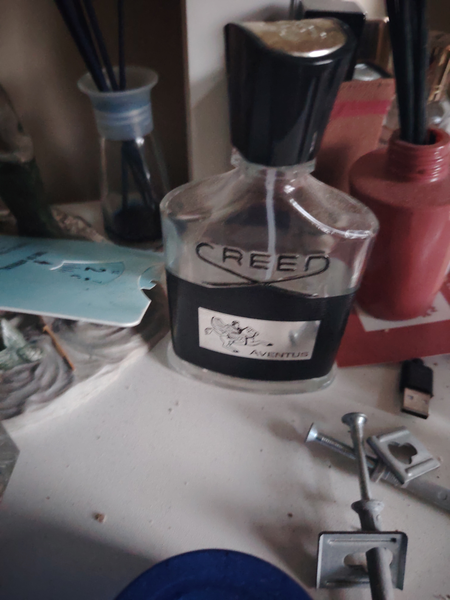 The image depicts an indoor scene with a cluttered, light-colored, possibly white or off-white, flat surface. Visible dust particles and small debris are present on the surface.
In the central foreground, a clear glass perfume bottle with a black, reflective cap is positioned. The bottle contains a dark liquid in its lower half, and a transparent spray tube is visible internally. The word "CREED" is embossed or printed near the top of the dark liquid line. A silver label adhered to the lower front of the bottle features an illustration of a figure on horseback and the word "AVENTUS" printed below it.
In the background, from left to right:
To the immediate left of the perfume bottle, a clear glass diffuser bottle with a light blue translucent stopper contains multiple dark diffuser reeds and a dark liquid.
Behind the perfume bottle, a reddish-brown rectangular object, possibly cardboard, is partially visible.
To the right of the perfume bottle, a reddish-orange cylindrical container, possibly a nail polish bottle or similar, has a dark stick protruding from its opening.
To the far right, a dark USB-A connector with a silver port is visible on the surface.
In the foreground, from left to right:
To the left, a light blue, irregularly shaped item, possibly a piece of plastic or paper, with faint dark markings (e.g., "23") is partially visible.
Further right, below the perfume bottle, a collection of metallic objects is arranged. These include one long, thin metal rod with a widened head, two shorter metallic pieces resembling screws or nails, and two 'butterfly' style drywall anchors.
In the bottom left corner, a segment of a dark blue circular object is visible.
No individuals or direct actions are depicted. The scene is static.
The image contains no discernible information to determine its geographical location, including any city within Huntingdonshire, United Kingdom.