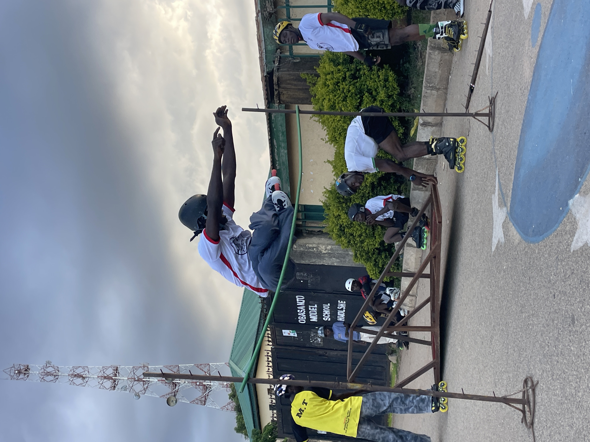 In Jos, Nigeria, a group of young men are engaged in rollerblading activities outside a school. The central figure is captured mid-air, performing a jump over a low horizontal bar set up with metal poles. This individual is wearing a helmet and a white shirt with red trim, along with grey pants and rollerblades. Several other individuals are present, some observing the jump, and others wearing helmets and rollerblades, suggesting they are part of the group or participants. In the background, a tall telecommunications tower stands against a cloudy sky, and the facade of the "OBASANJO MODEL SCHOOL HIADLSHE" is visible. The scene appears to be taking place on a paved surface, possibly a road or court, with some greenery and fencing in the surroundings.