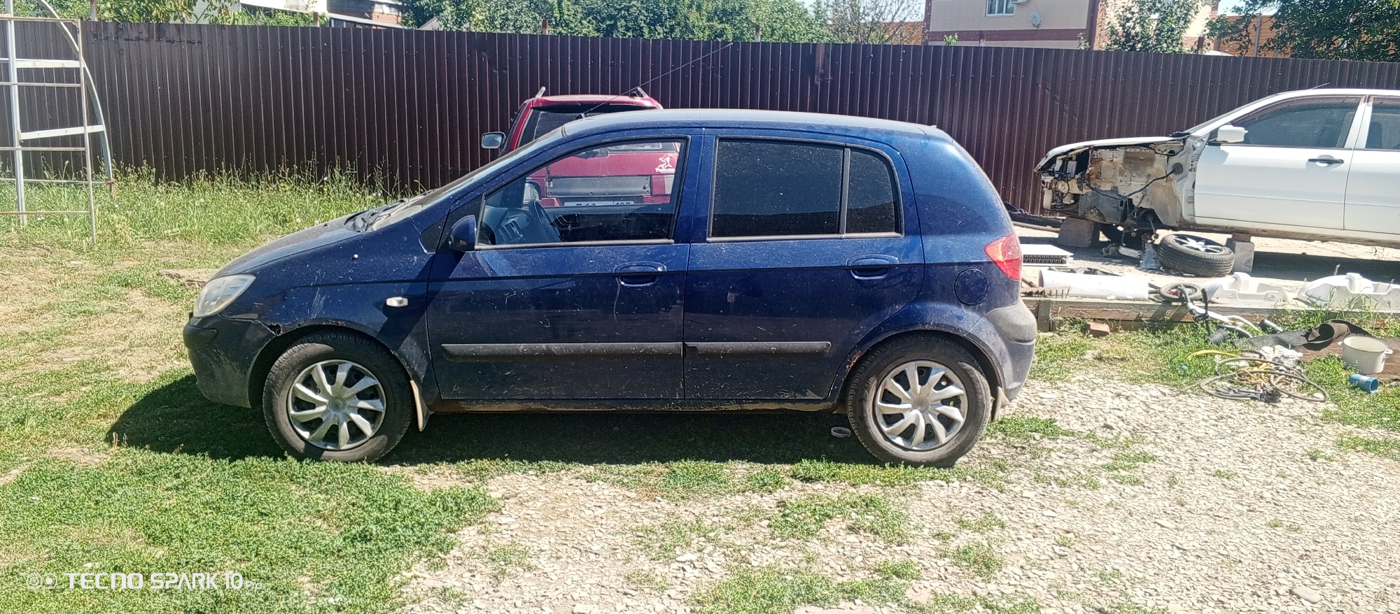 The image shows a blue four-door hatchback car parked outdoors on a sunny day. The car is dirty and appears to be in a state of disrepair, with visible scuffs and dirt on its exterior. It is parked on a surface that is a mix of gravel and grass. In the background, a brown metal fence runs along the property line, with some trees and a building visible beyond it. To the right of the hatchback, a white car is partially visible, appearing to be dismantled or heavily damaged, with its front end removed. Various car parts and tools are scattered on the ground around the white car, suggesting a possible auto repair or salvage yard. A red SUV is also visible behind the blue hatchback, partially obscured by it. The location is identified as Bereg Kubani, Russia.