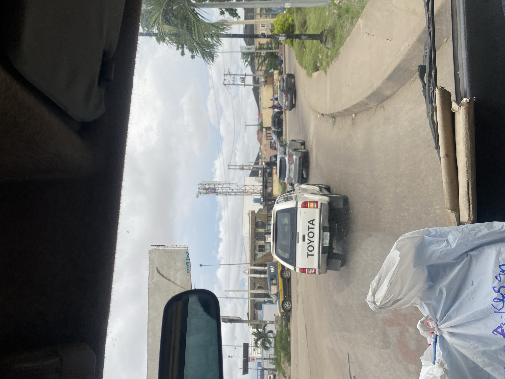This image, captured from inside a vehicle in Ikeja, Nigeria, shows a street scene with several cars. In the foreground, a white Toyota pickup truck with the license plate "APP 2910U" is visible from the rear. To the left, a blue and yellow car is parked. In the middle ground, a silver SUV is driving away from the camera, and a dark sedan is also on the road. Several people can be seen walking in the background. The environment includes buildings, palm trees, and utility poles. The sky is overcast with clouds.