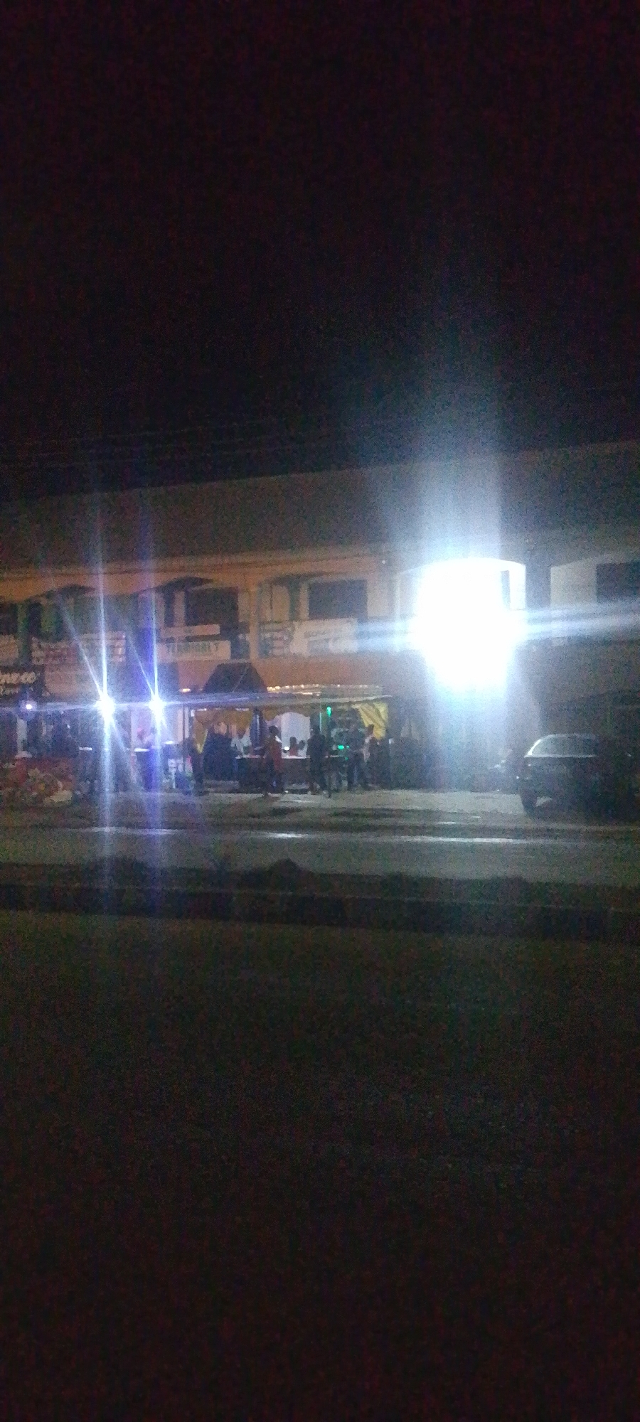 The image depicts an outdoor scene at night, likely a street or commercial area in Gura Top, Nigeria. A building with a slightly raised awning or roof structure is visible, illuminated by bright, harsh lights, some of which create lens flare effects. People are gathered in front of the building, suggesting a social event or a marketplace. Several vehicles, including a dark-colored car on the right side of the image, are present. The foreground shows a grassy area, possibly a median or parkway, separated by a darker, lower strip that could be a curb or a drainage ditch. The overall atmosphere is dimly lit, with the artificial lights being the primary source of illumination.
