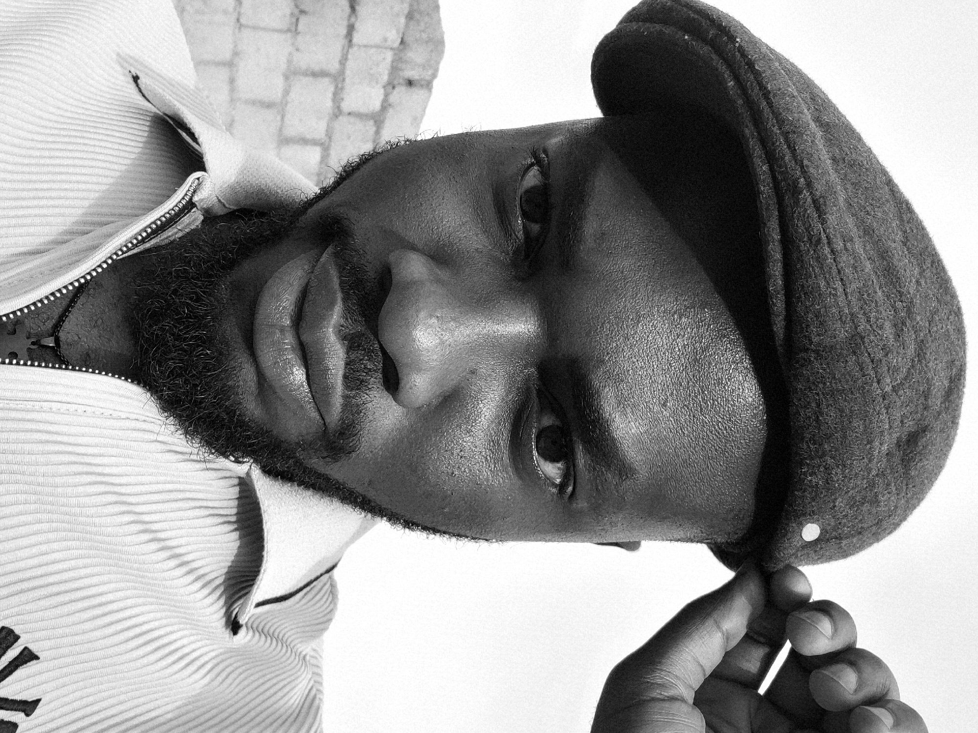 A close-up, black and white portrait shows a Black man wearing a newsboy cap. He is facing the viewer with his head tilted to the side. He has a beard and his lips appear slightly parted. He is wearing a striped shirt with a zipper. His fingers are touching the rim of the cap. Behind him, there is a blurred background showing some bricks. There is no explicit evidence to confirm this image was taken in Jos, Nigeria.