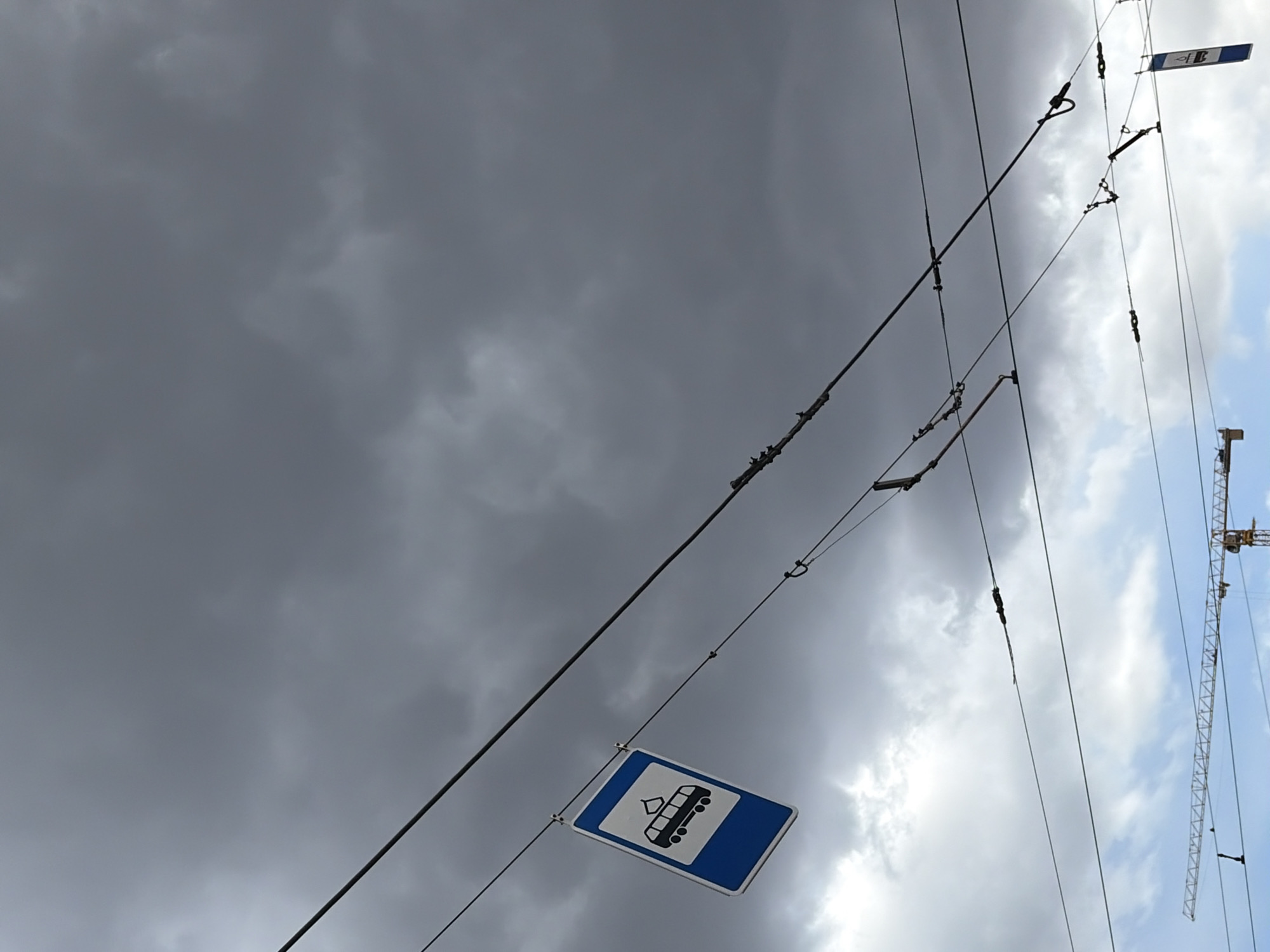 The image depicts a view towards the sky in what appears to be the city of Perm, Russia. Overhead electrical lines are strung against a backdrop of cloudy sky, with some dark storm clouds present. A sign, blue and white, showing an image of a trolleybus hangs from one of the overhead lines. In the background, a construction crane is visible on the right side of the image.