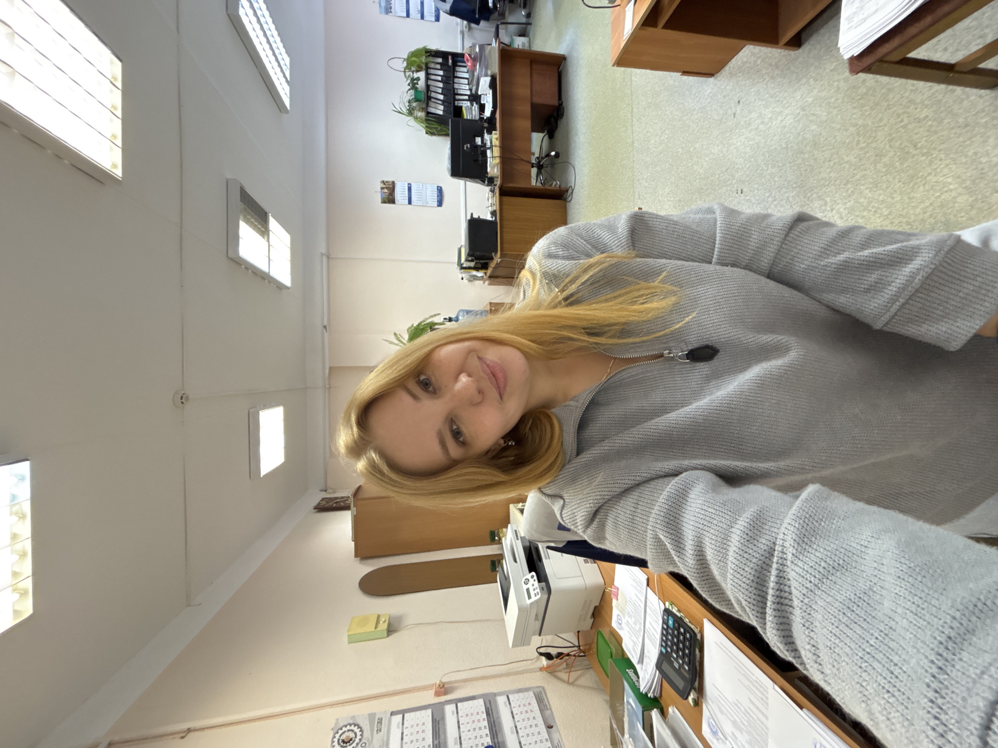 The image shows a woman with blonde hair wearing a gray sweater in an office setting. The office has fluorescent lights, desks, computers, a printer, calendars, and office supplies. The location is Unknown, Unknown.