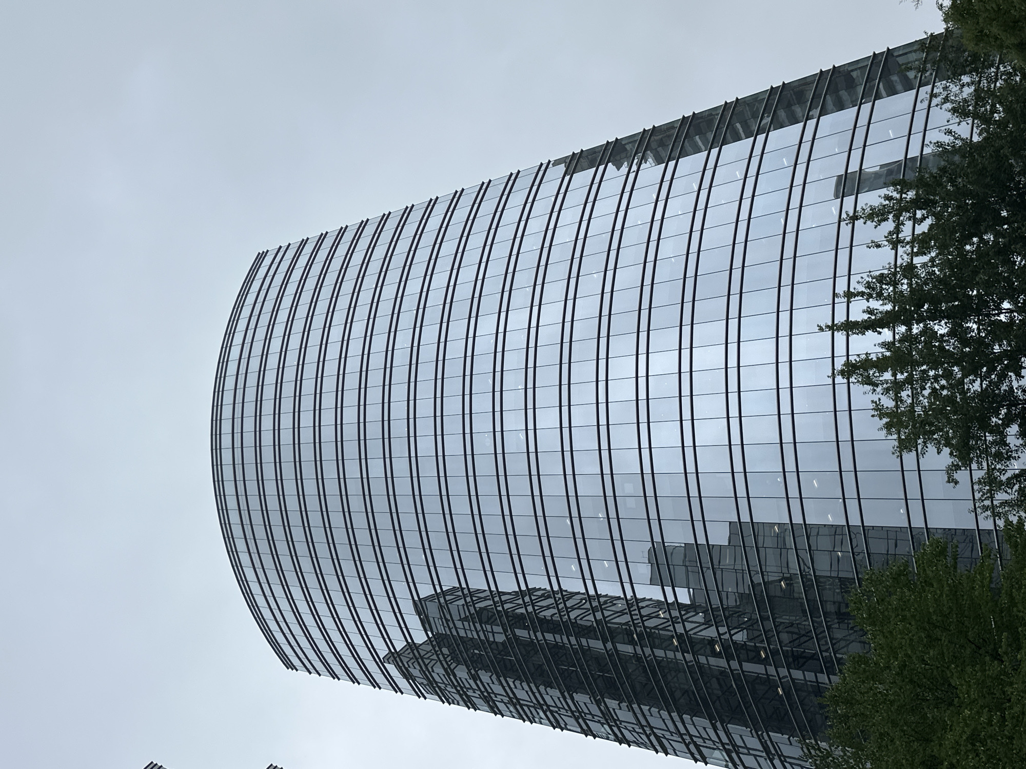 A low-angle, outdoor, full-shot photograph captures a modern, curved skyscraper in Burnaby, Canada, against an overcast sky. The building is constructed with reflective glass panels that form horizontal bands, giving it a layered appearance. The glass reflects the sky and another, taller skyscraper, which is visible as a dark, angular shape on the left side of the frame. The lower portion of the image features the tops of lush green trees, obscuring the base of the skyscraper. The overall scene conveys a sense of urban architecture and a cloudy day.