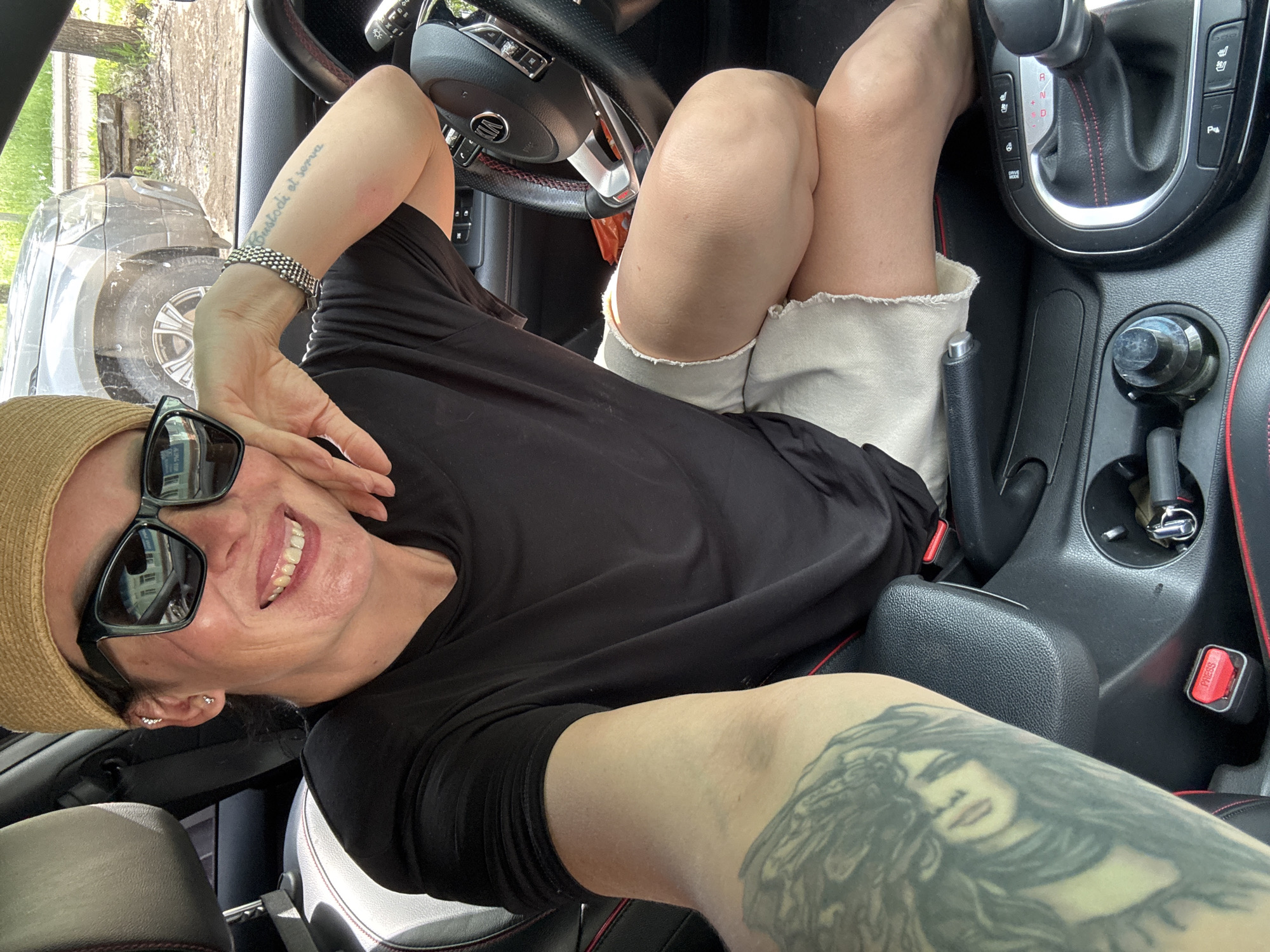 A woman with dark hair wearing a straw hat, sunglasses, a black t-shirt, and light-colored shorts is sitting in the driver's seat of a car. She is smiling and resting her chin on her hand. The interior of the car, including the steering wheel, dashboard, and gear shift, is visible. The woman has a tattoo on her left forearm and a bracelet on her right wrist. The car appears to be parked outdoors, with another vehicle visible through the windshield. The image is a selfie taken by the woman. The specific location in the city of Chaikovsky, Russia cannot be determined from the image alone.