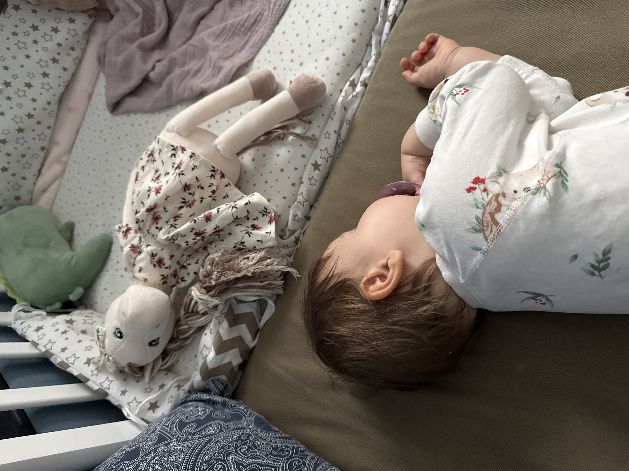 A baby is sleeping on a brown surface next to a crib. The baby is wearing a white onesie with deer prints and has a pacifier in its mouth. The crib contains a green dinosaur plush toy, a doll with a floral dress and fringe hair, bedding with star patterns, and a lavender cloth. A blue paisley cushion is visible at the bottom of the crib. The location cannot be determined from the image, so there is no proof this image was taken in Moscow, Russia.