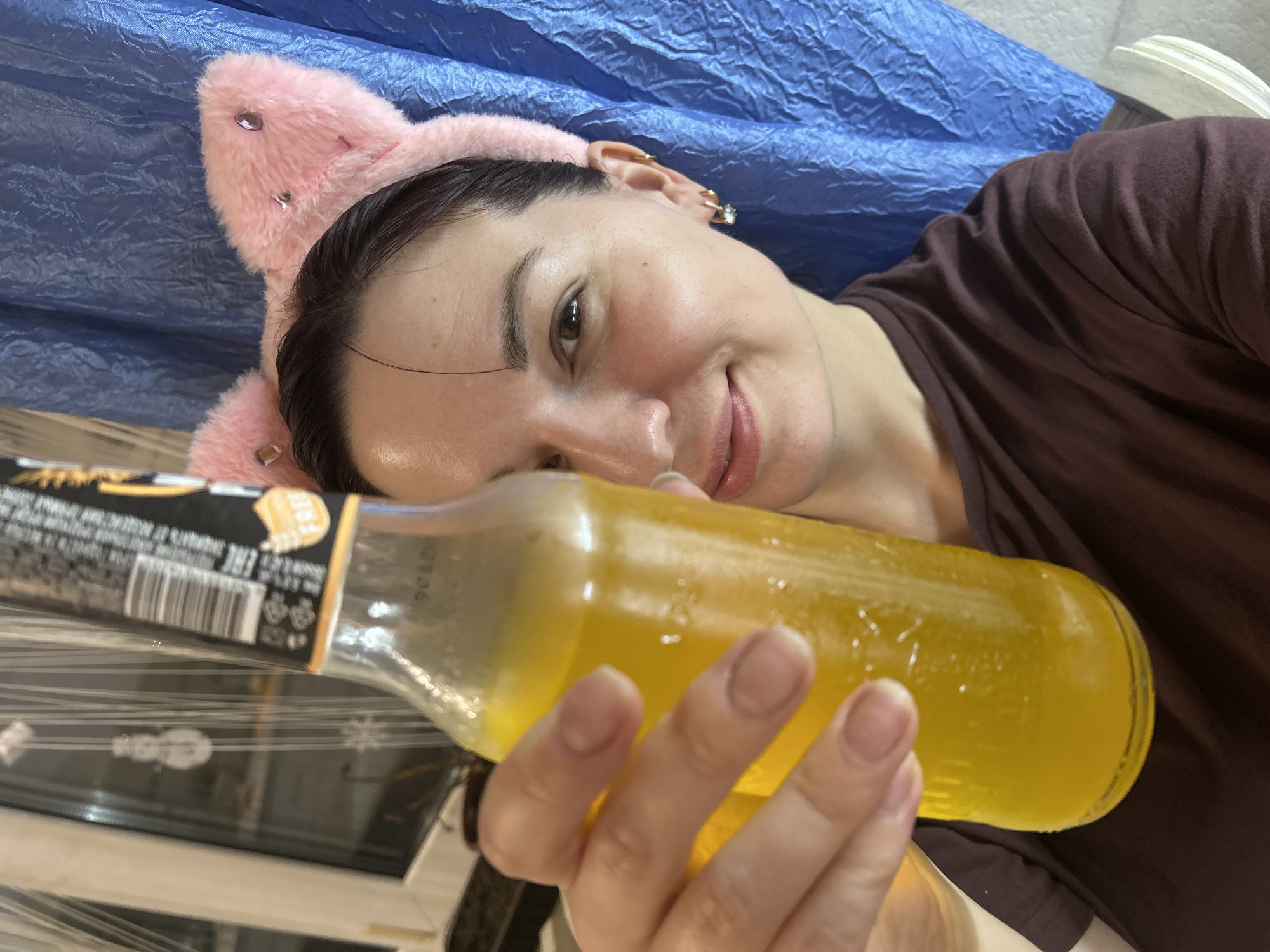 A young woman with dark hair and a playful pink cat-ear headband is holding up a glass bottle of yellow liquid towards the camera. She is wearing a dark maroon t-shirt and has on small hoop earrings. She is smiling at the camera. Behind her, a blue tarp is draped, and to the left, a window with a white frame is visible. A subtle decoration resembling a snowman hangs on the window. The scene appears to be indoors, possibly a room in a home. The overall atmosphere suggests a casual and lighthearted moment. The location is identified as Фоки, Russia.